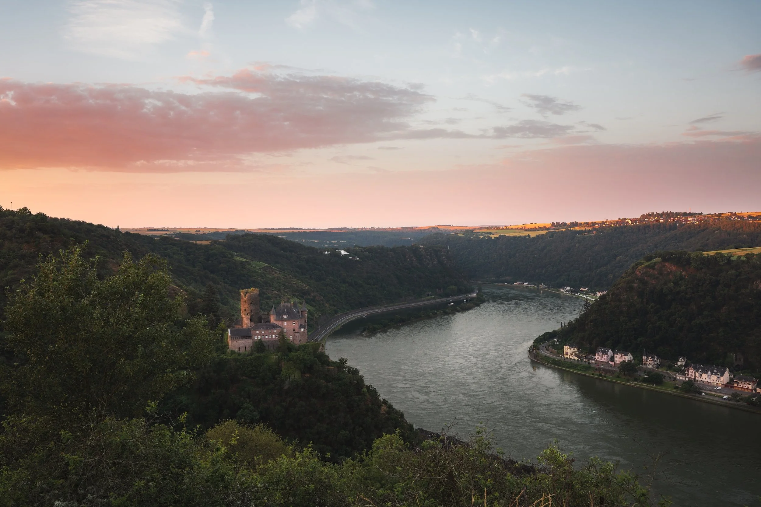 Ein Fluss fließt durch eine bewaldete Landschaft bei Sonnenaufgang, mit einem mittelalterlichen Schloss auf einem Hügel und kleinen Häusern am Flussufer.