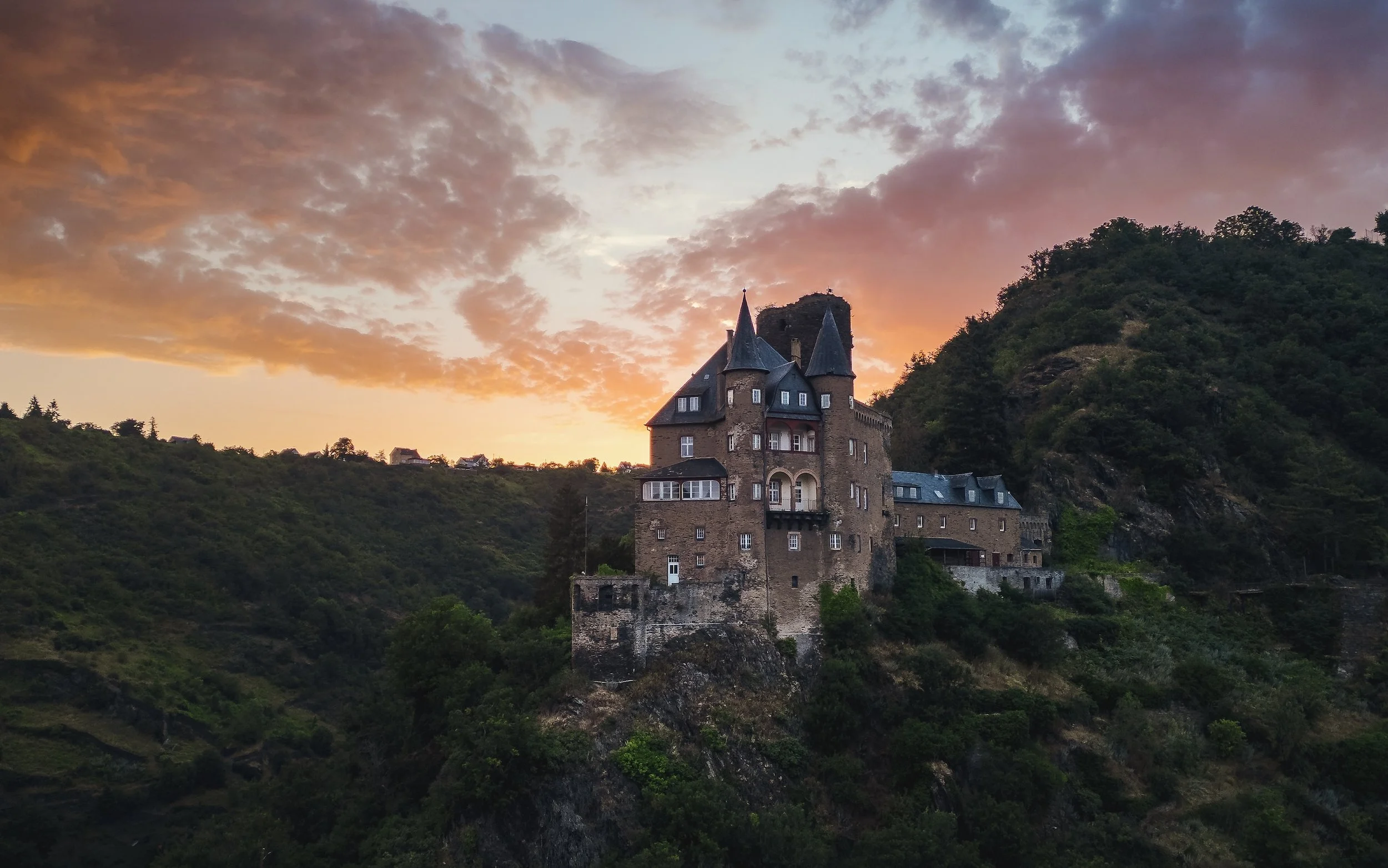 Mittelalterliches Schloss auf einem Hügel bei Sonnenuntergang, umgeben von grünen Hügeln und bewölktem Himmel.