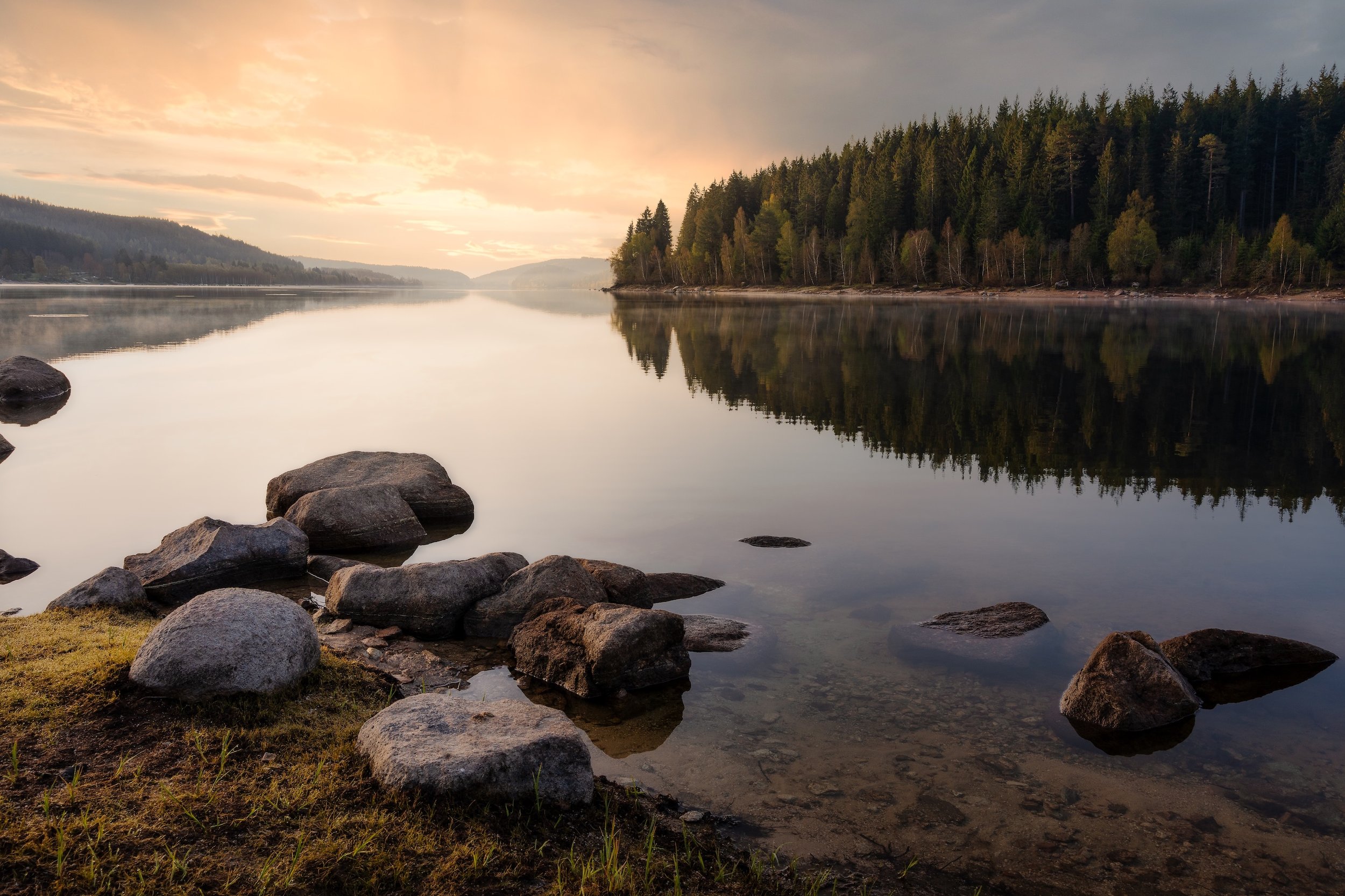 Ein ruhiger See mit Steinen am Ufer, umgeben von einem dichten Wald, bei Sonnenuntergang.
