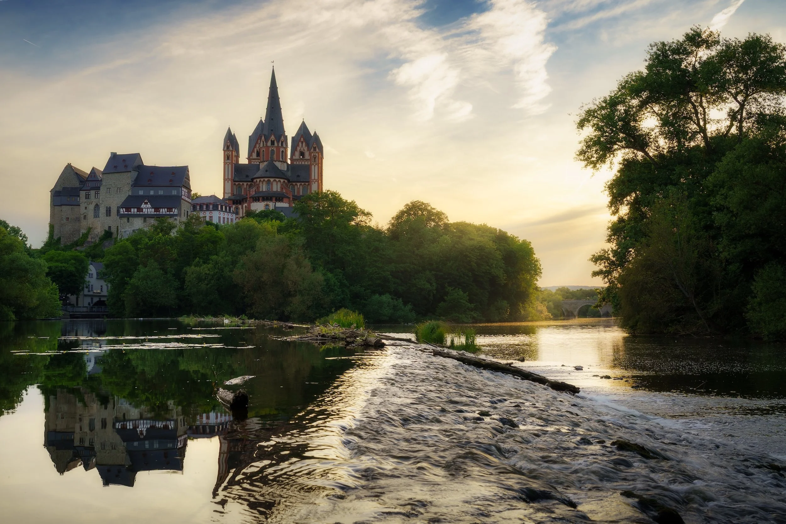Burg auf einem Hügel neben einem Fluss bei Sonnenuntergang, umgeben von Bäumen.