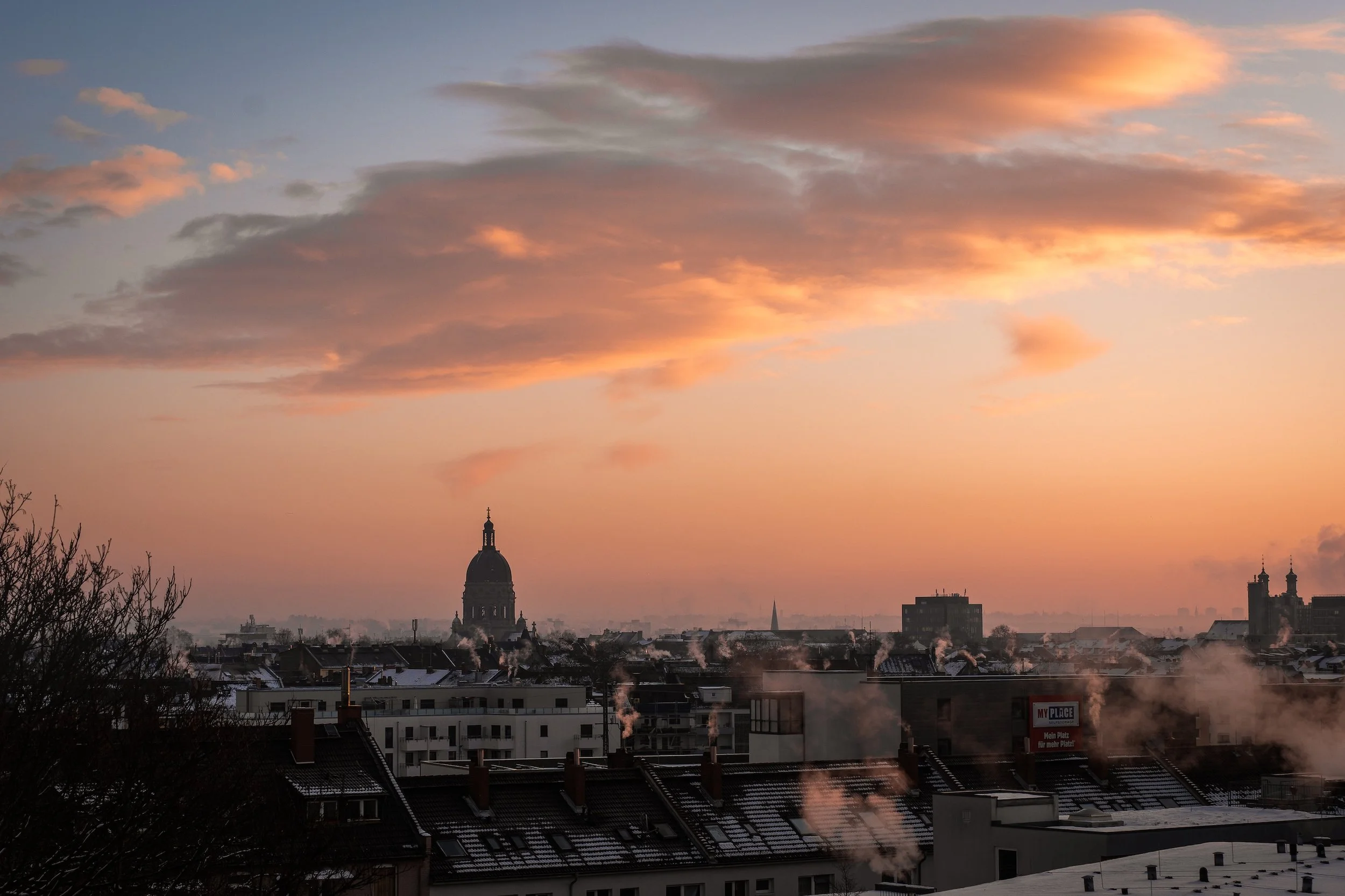 Blick auf eine Stadt bei Sonnenuntergang mit Wolken am Himmel, mehreren Rauchschwaden aus den Schornsteinen, einem historischen Dome im Hintergrund und Schneedeckten Dächern im Vordergrund.