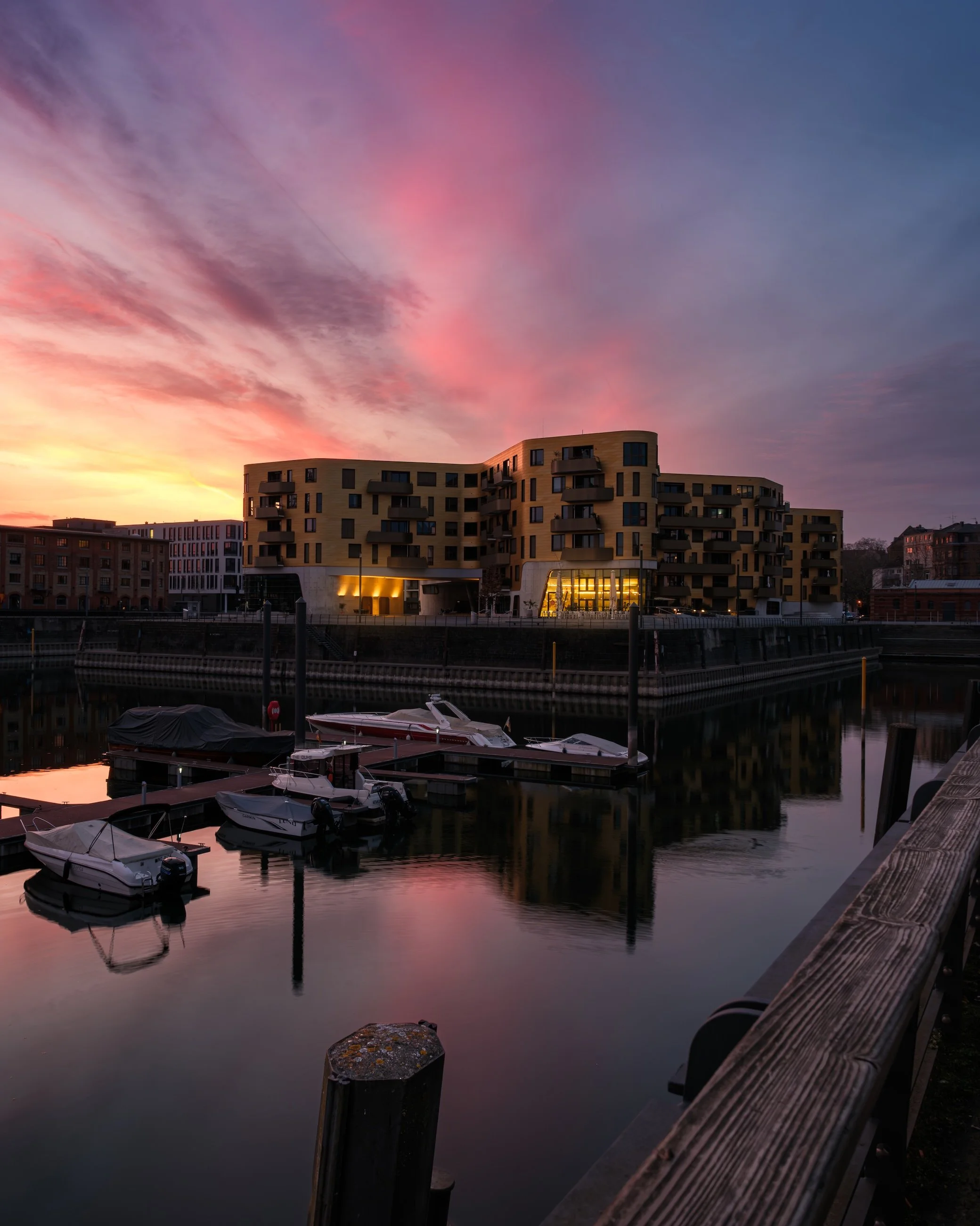 Ein moderner Gebäudekomplex am Wasser bei Sonnenuntergang mit reflektierendem Himmel und Booten im Vordergrund.