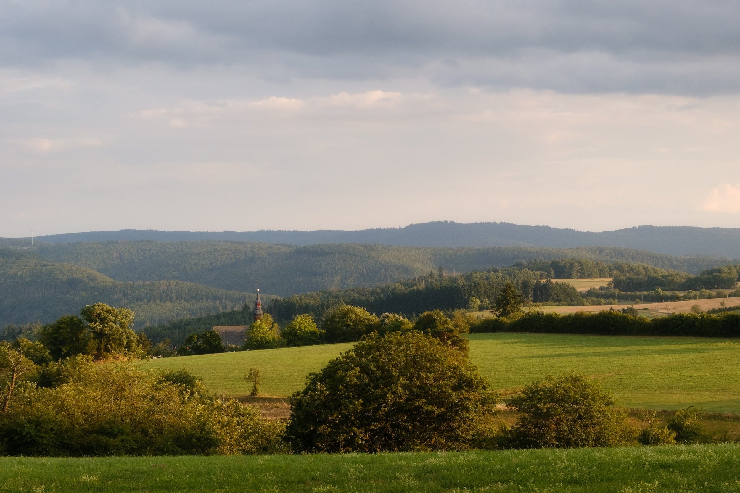 Landschaft mit grünen Wiesen, Bäumen und Hügeln, entfernter Kirchturm, bewölkter Himmel.