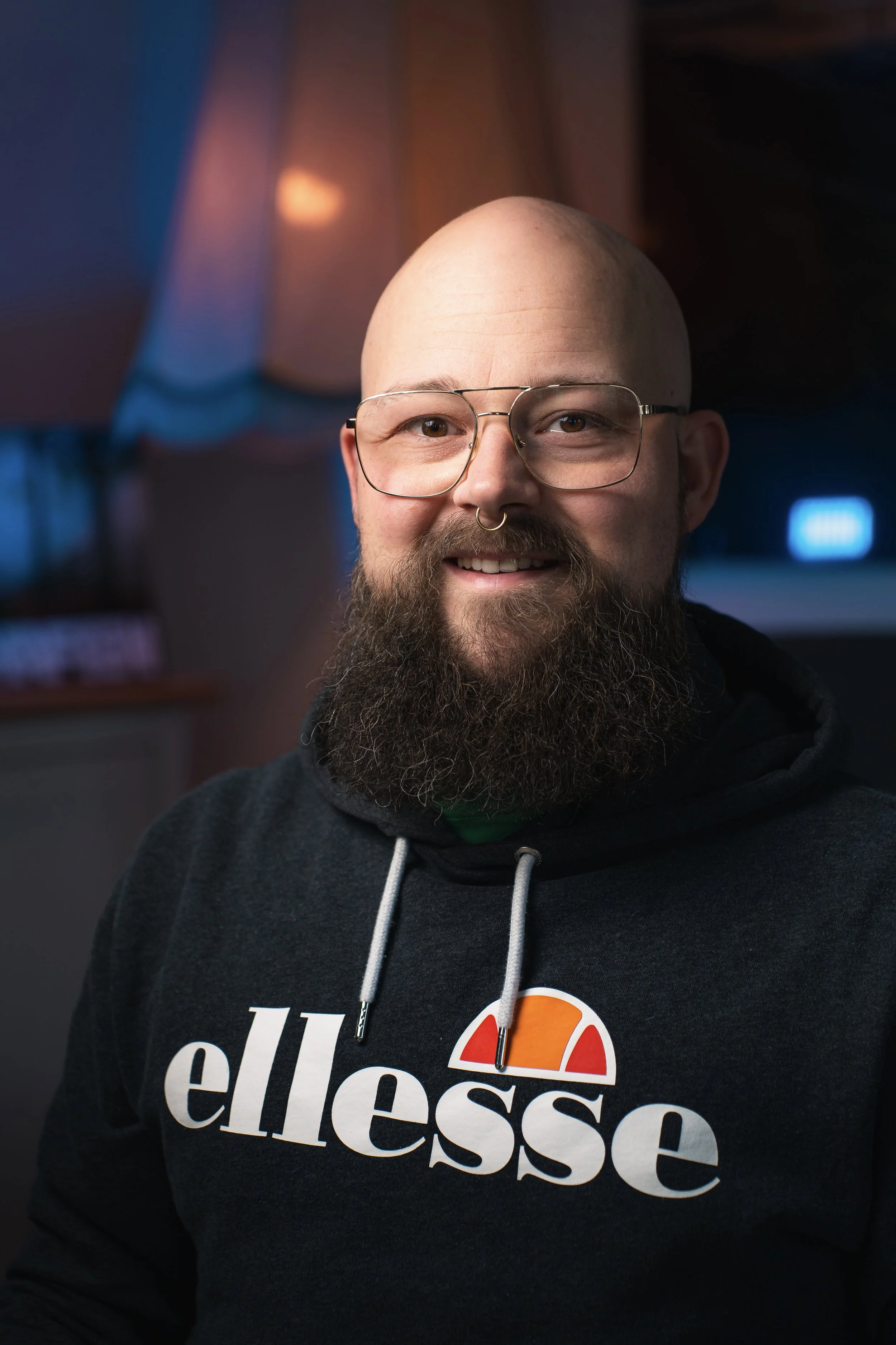 Ein lächelnder Mann mit Brille, Bart und Nasenpiercing trägt ein schwarzes Ellesse Hoodie, im Hintergrund sind bunte Lichter in einem dunklen Raum sichtbar.