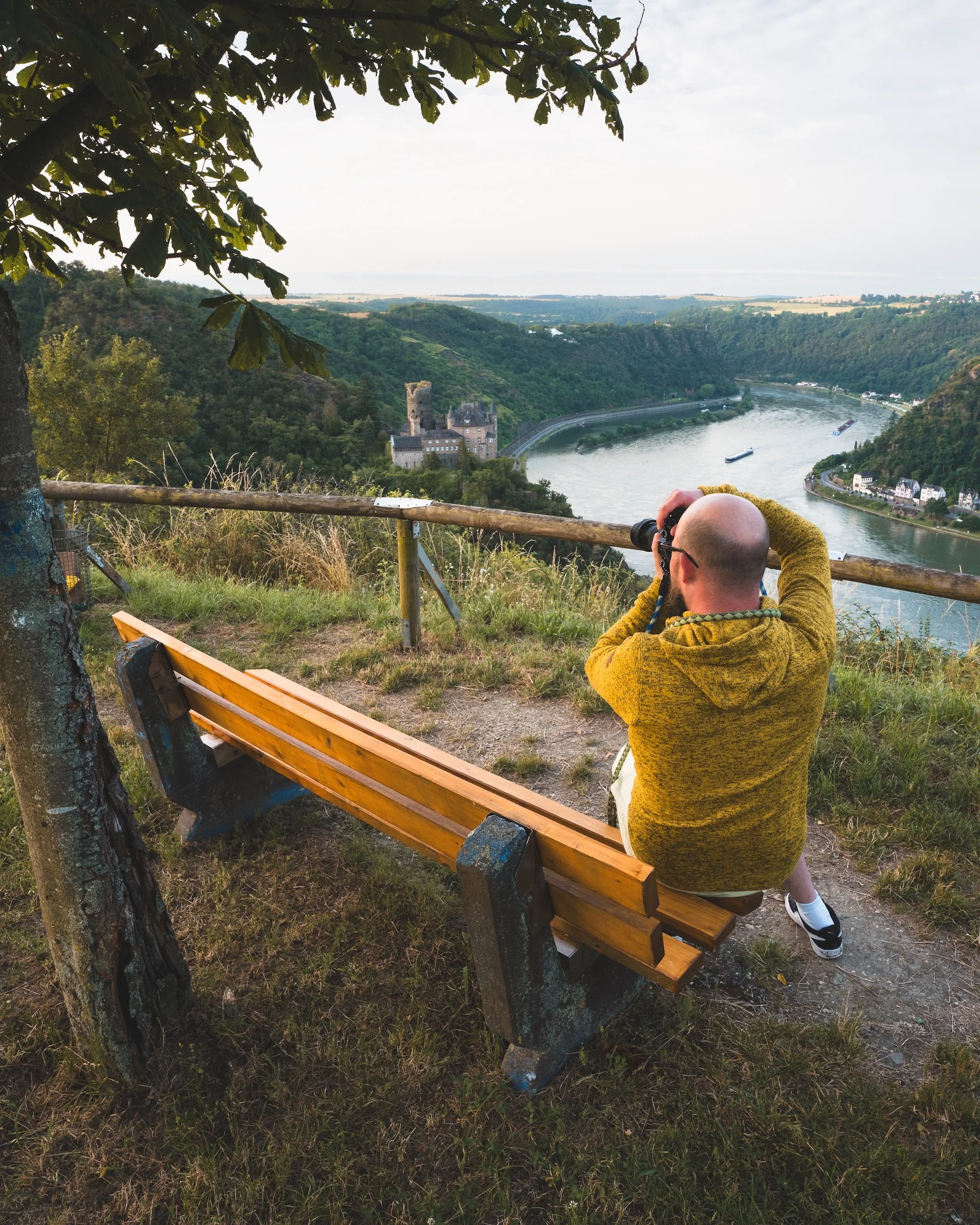 Ein Mann sitzt auf einer Bank auf einem Hügel und schaut durch ein Fernglas auf einen Fluss, umgeben von grünen Hügeln und einer Burg in der Ferne.