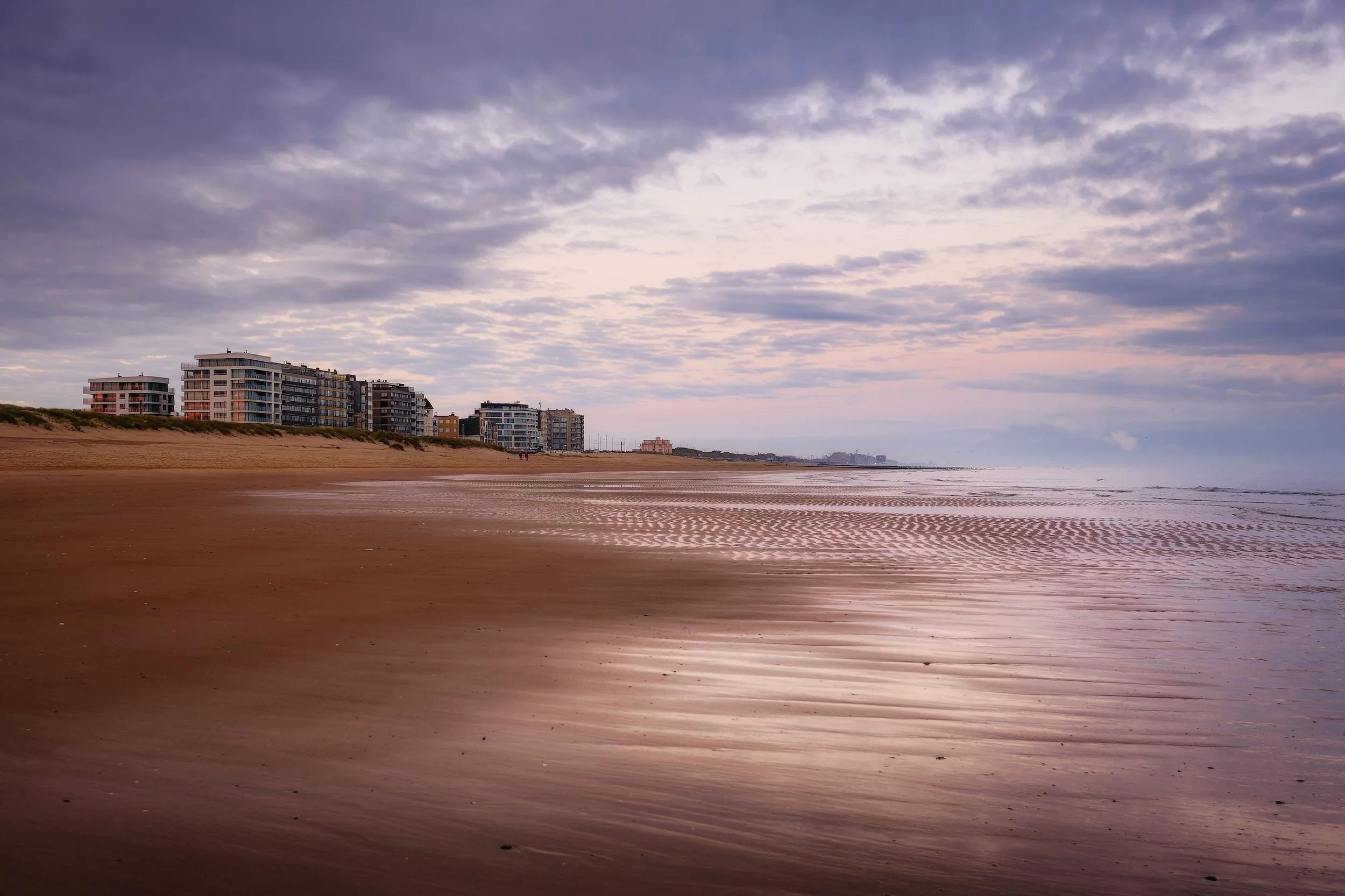 Leerer Strand bei Sonnenaufgang mit Gebäuden im Hintergrund und bewölktem Himmel.