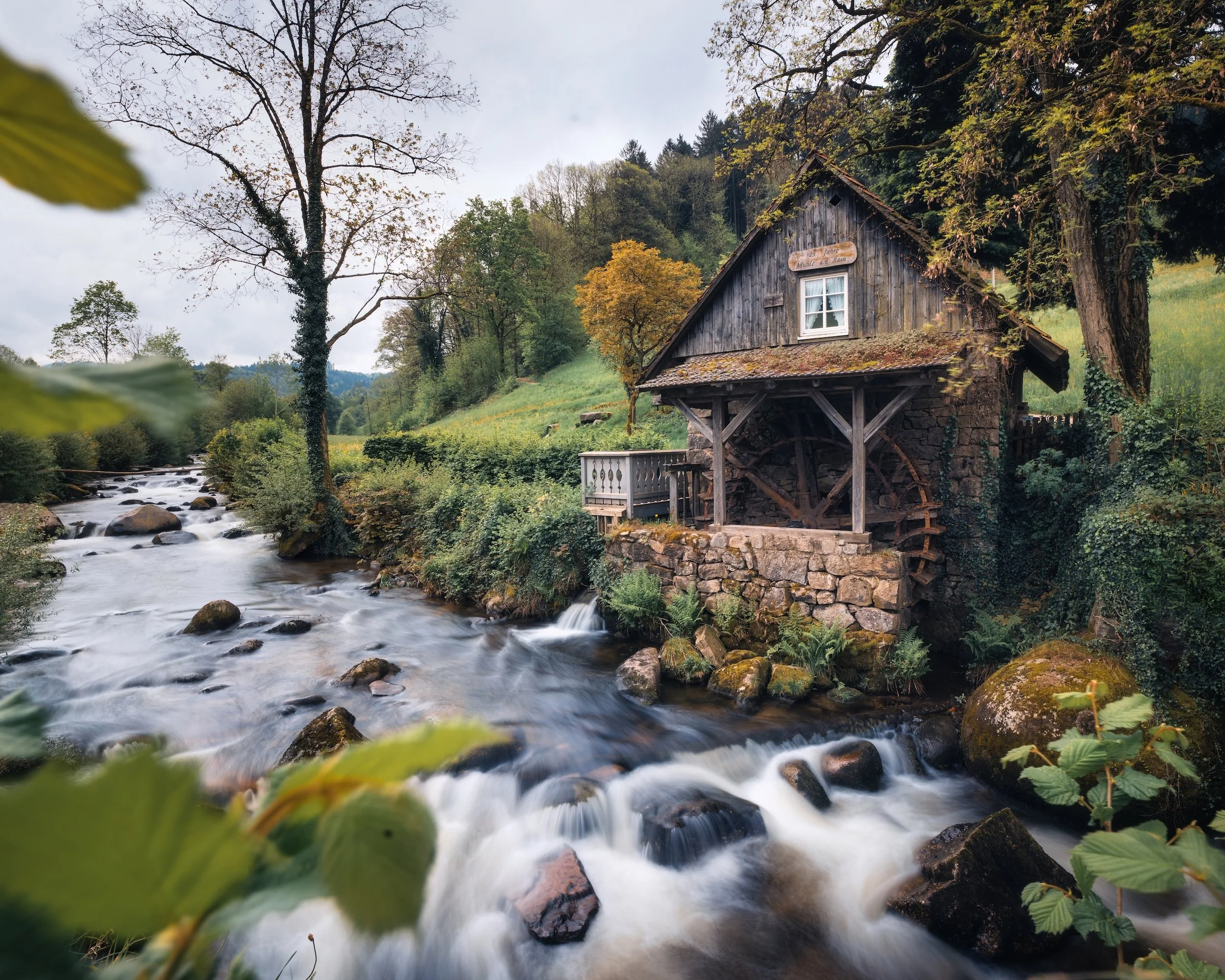 Ein alter Wassermühlen-Gebäude neben einem fließenden Fluss, umgeben von Bäumen und grünem Landschaft in einer ländlichen Gegend.