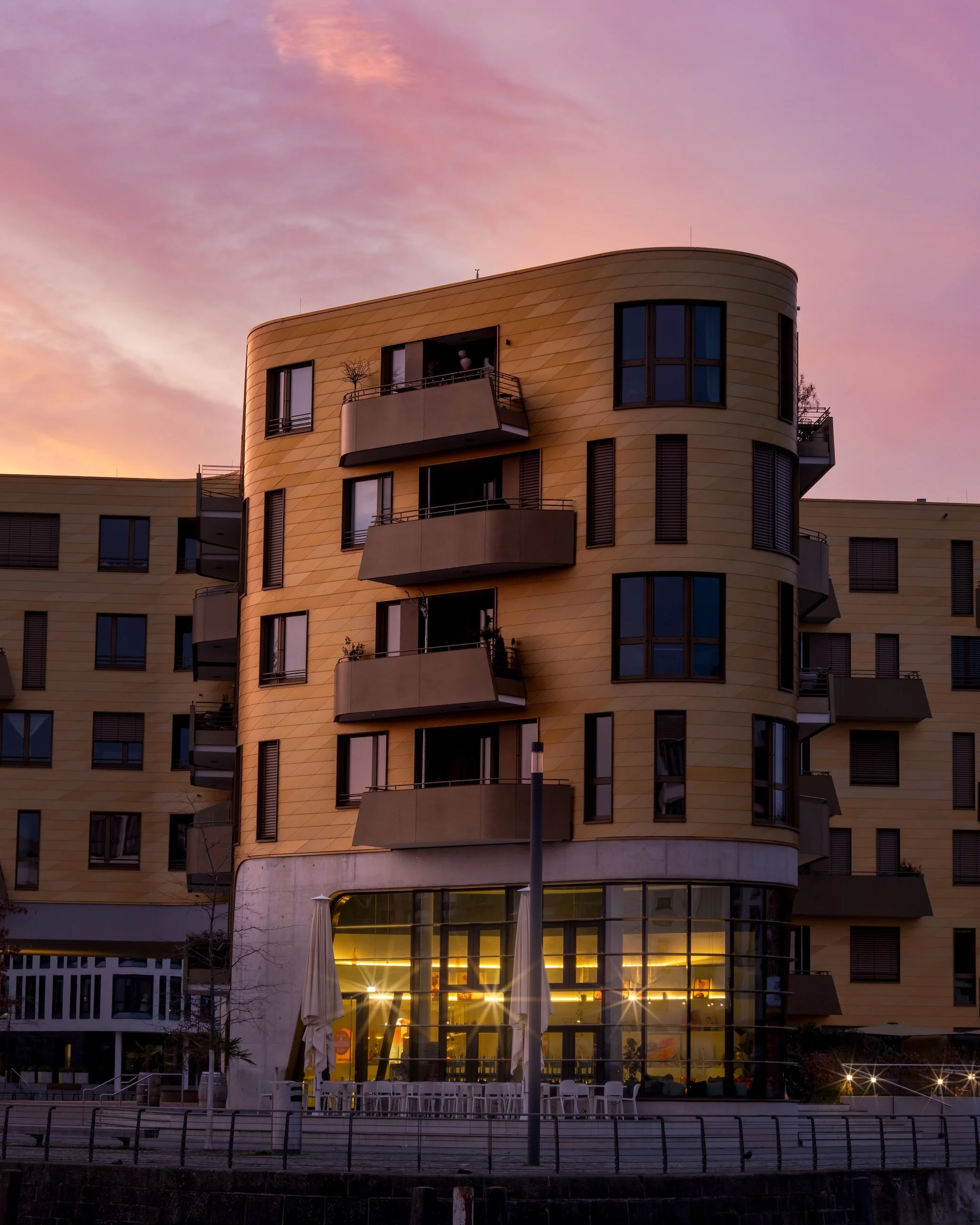 Modernes, gelbes Wohngebäude mit mehreren Balkonen bei Sonnenuntergang, große Fenster im Erdgeschoss mit Innenbeleuchtung.