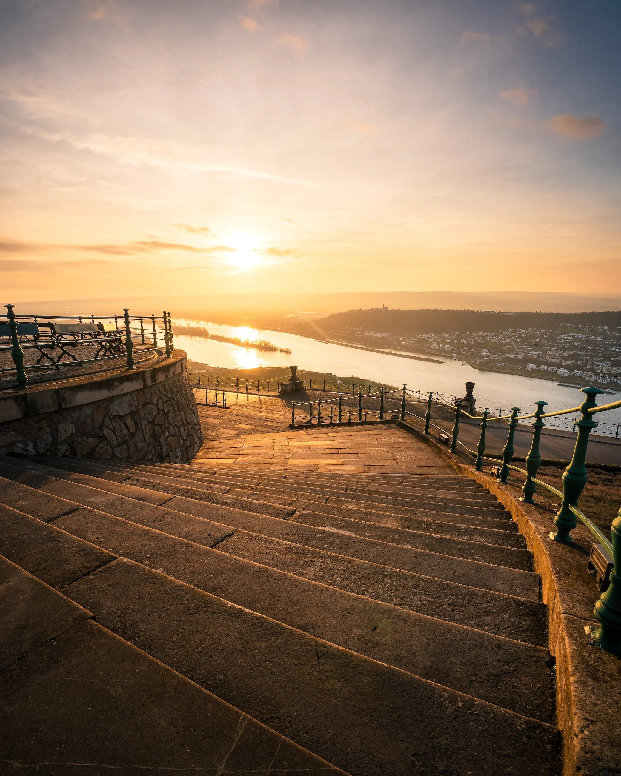 A stone staircase leading down to a riverside promenade, with a decorative railing, overlooking a river at sunset.