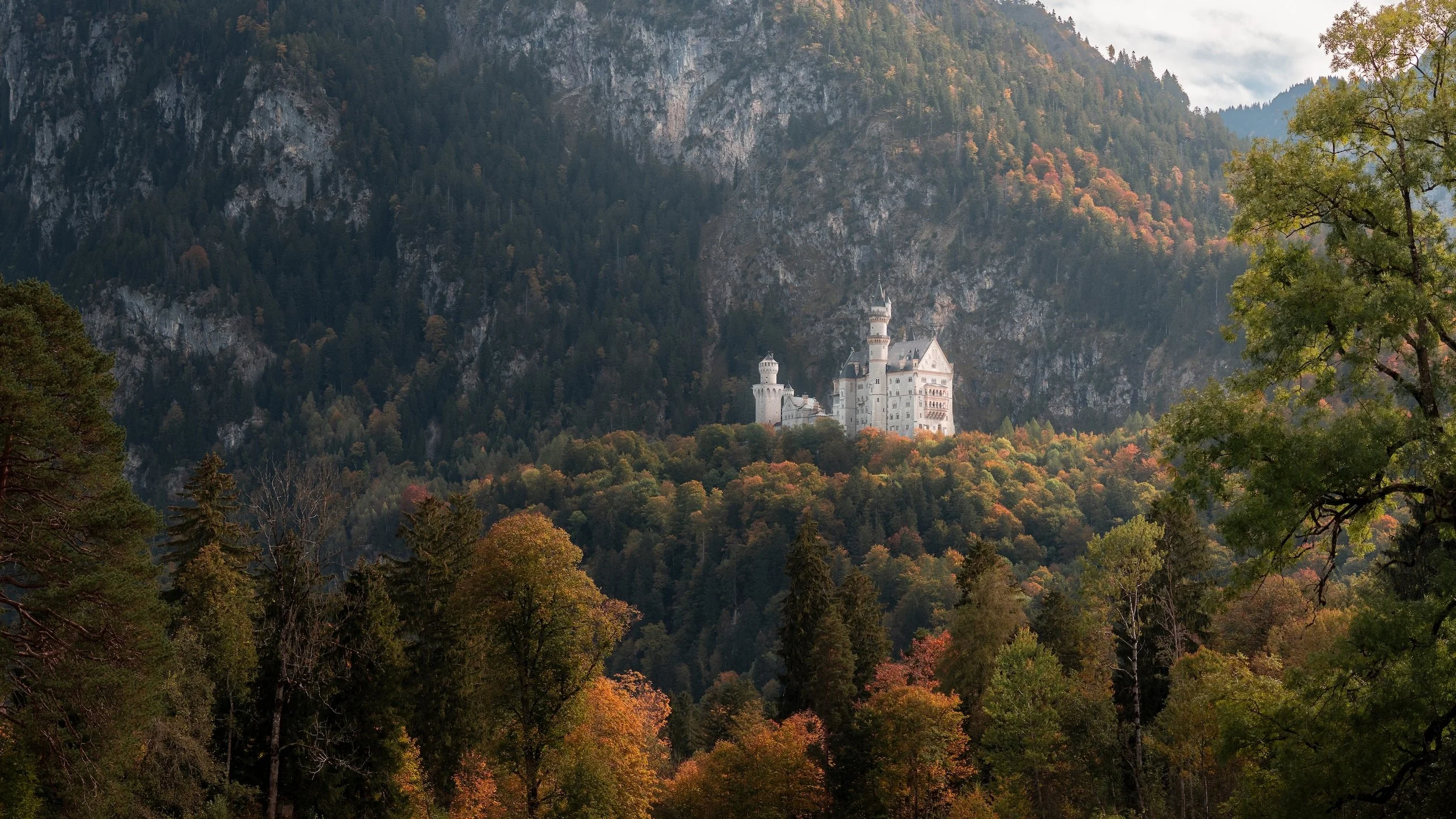Mittelalterliches Schloss auf einem Hügel inmitten eines dichten, herbstlich gefärbten Waldes, umgeben von hohen Bergen.