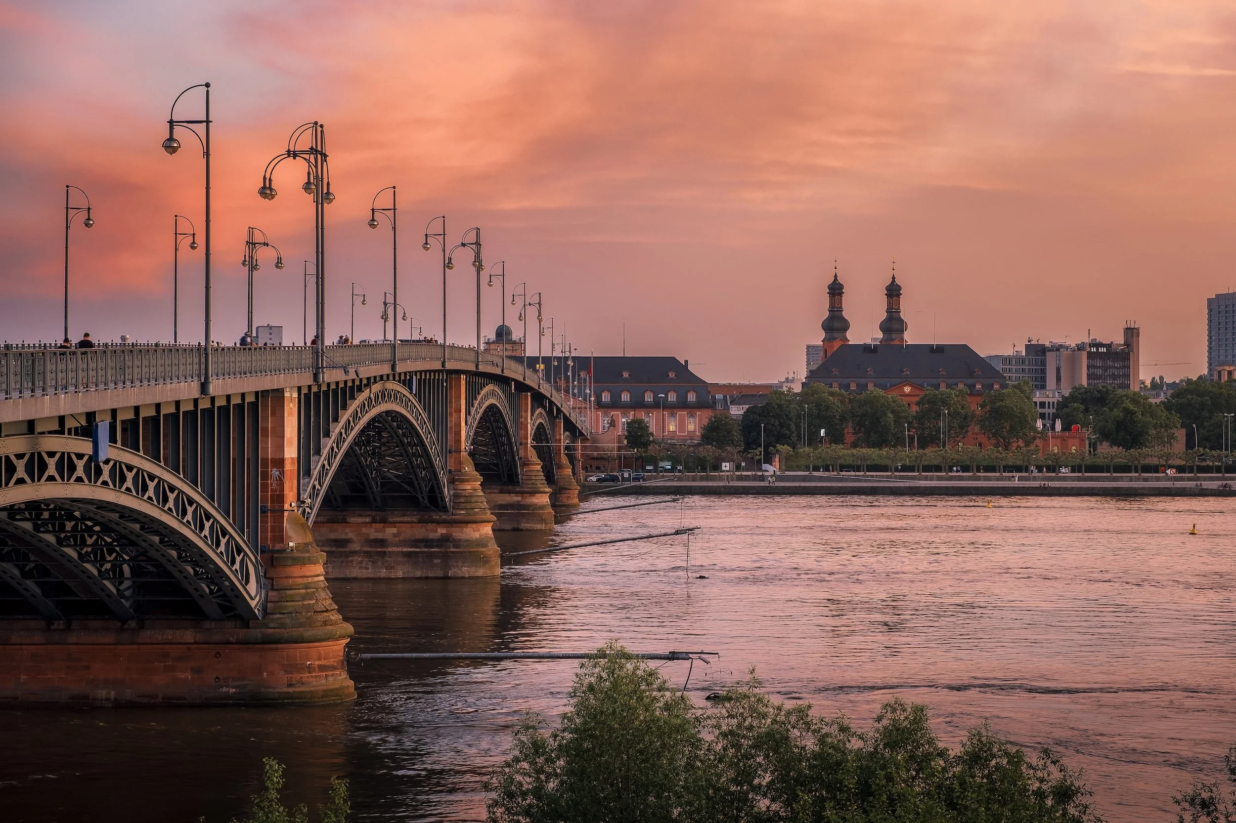 Ein Blick auf eine Brücke über den Fluss bei Sonnenuntergang, mit mehreren Lampen entlang der Brücke und einem städtischen Hintergrund mit Kirchen und Gebäuden.