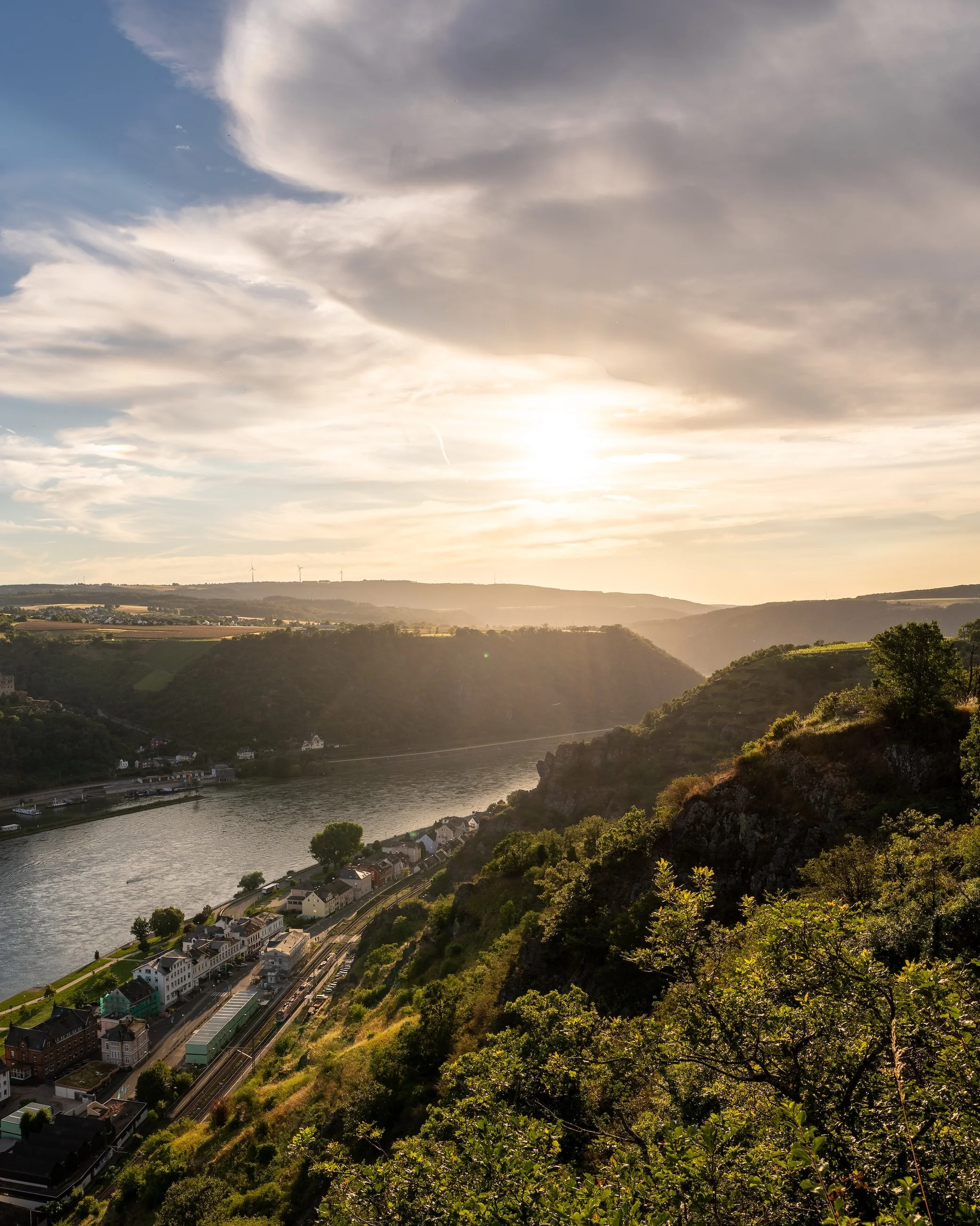 Landschaft mit Fluss, Hügeln, Bäumen und Sonnenuntergang am Himmel.