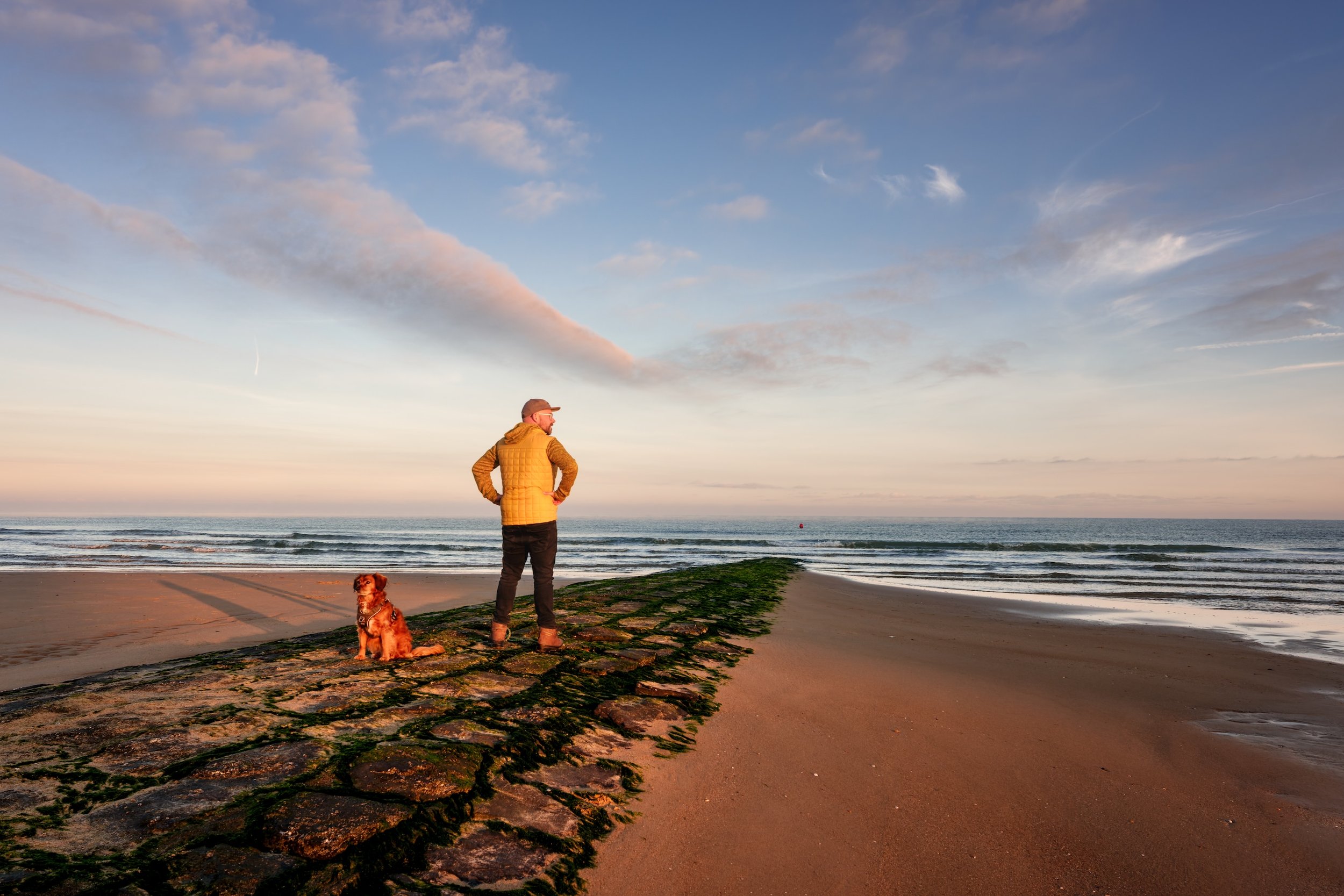 Ein Mann mit Hund steht auf einem Steg am Strand und schaut aufs Meer bei Sonnenuntergang.
