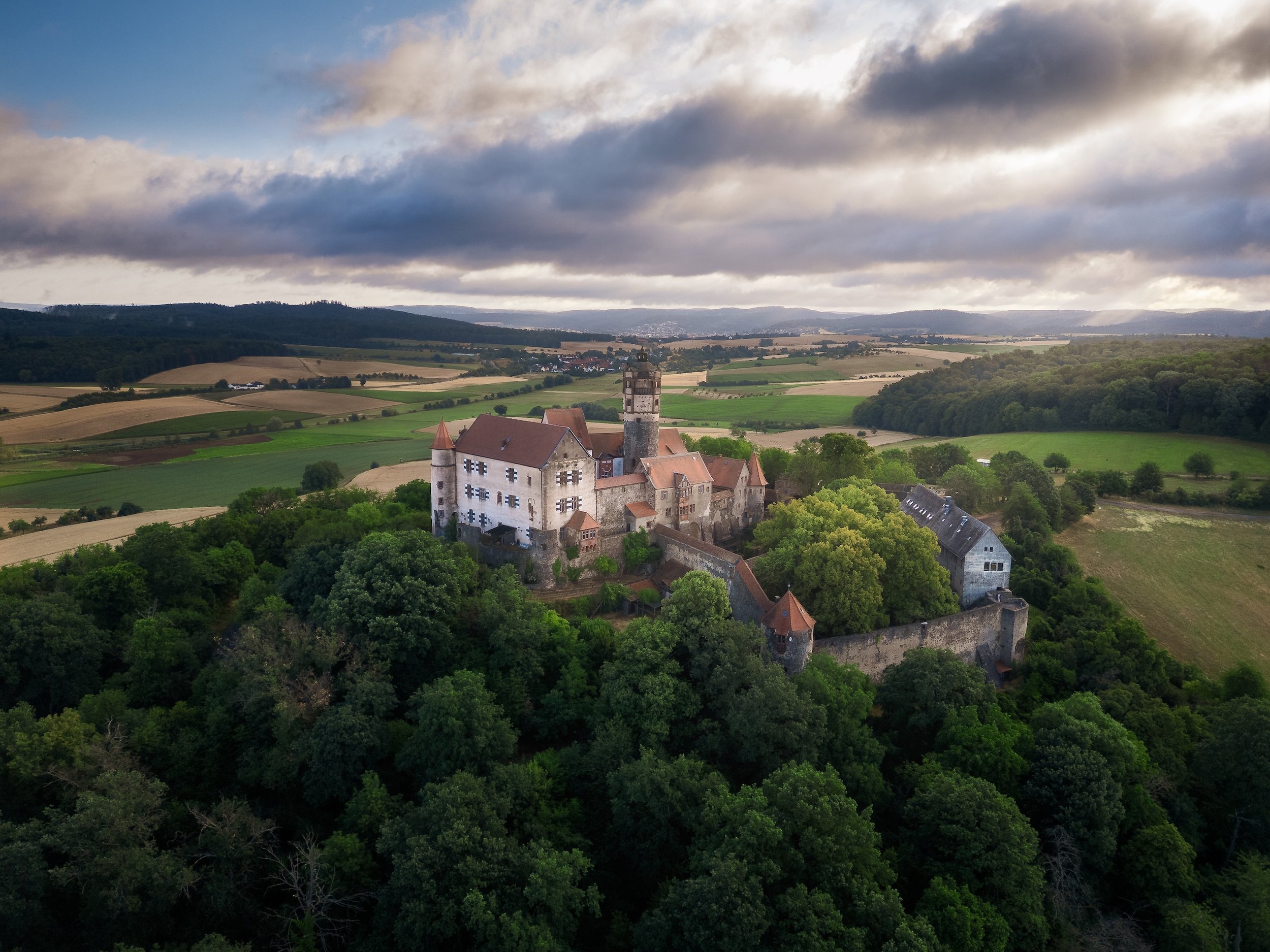 Ein mittelalterliches Schloss auf einem Hügel, umgeben von grünen Bäumen mit einer weiten Landschaft im Hintergrund, unter einem bewölkten Himmel.