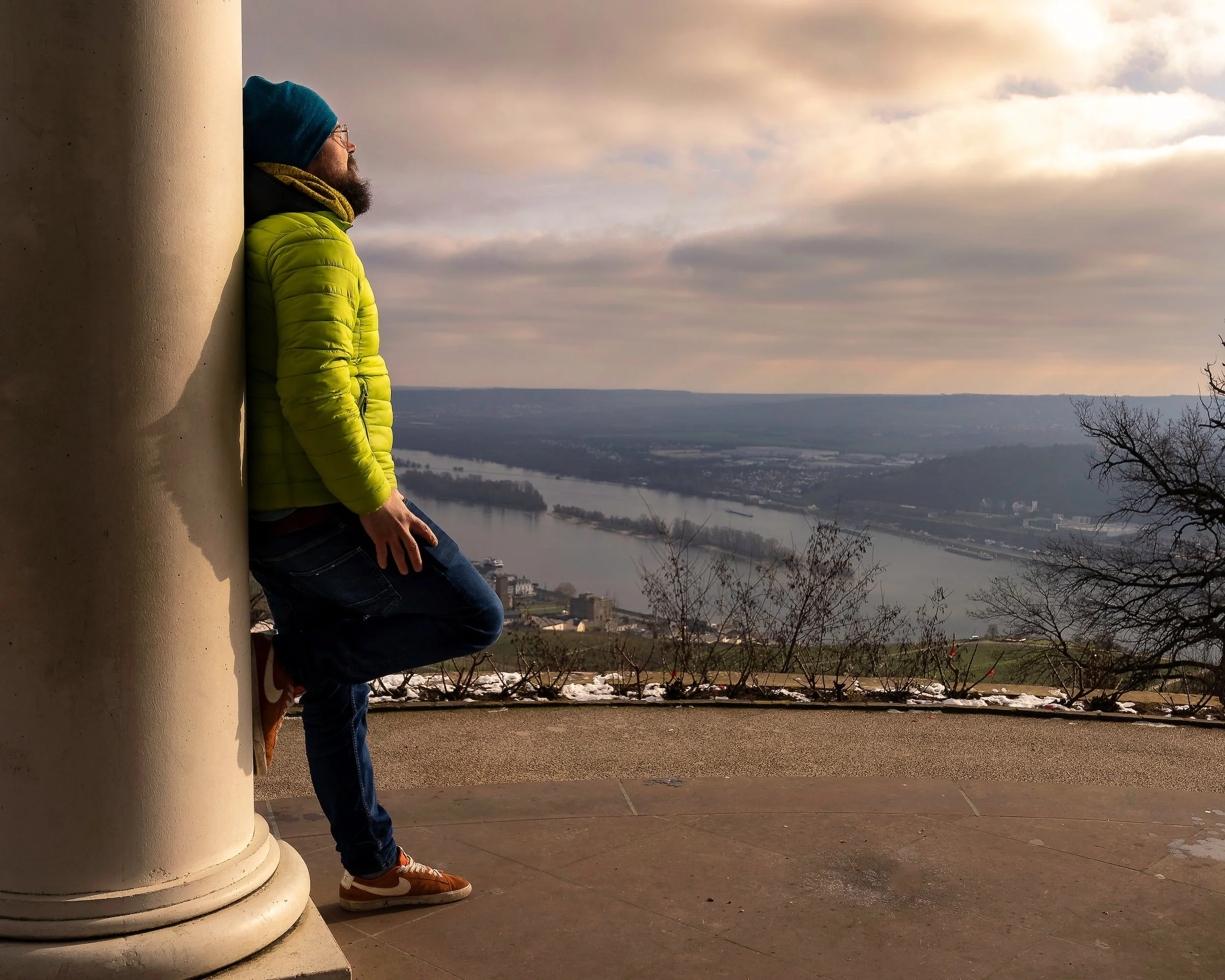 Ein Mann lehnt an einer großen Säule und schaut auf den Fluss und die Landschaft im Hintergrund, bei bewölktem Himmel.