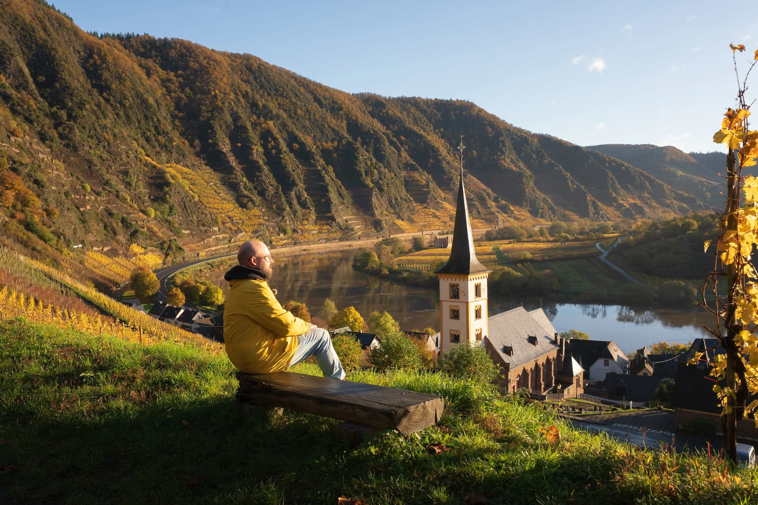 Ein Mann sitzt auf einer Holzbank und blickt auf eine malerische Landschaft mit einem Fluss, einem Kirchturm und bewaldeten Hügeln im Herbst.