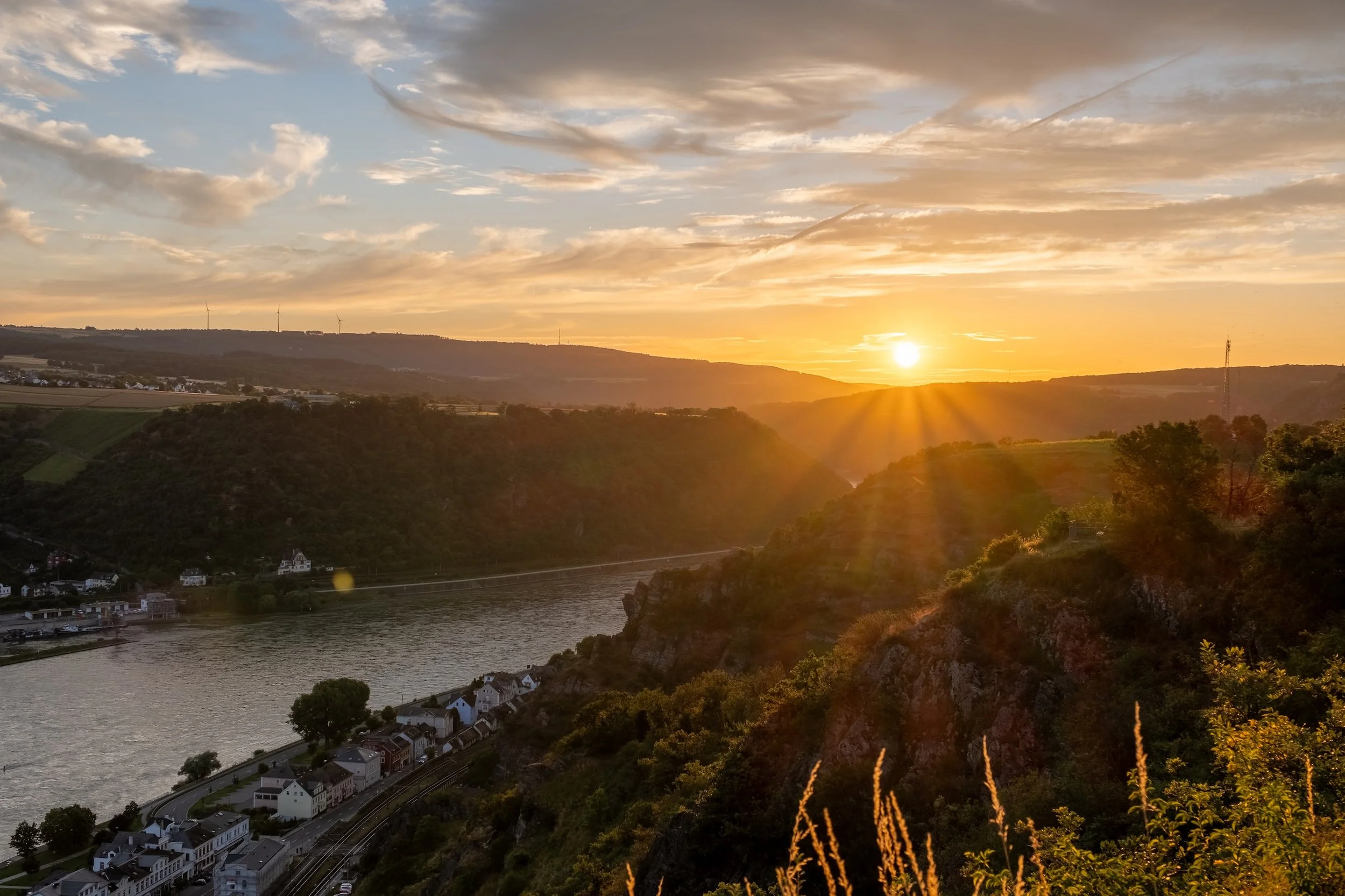 Blick auf einen Fluss bei Sonnenuntergang, umgeben von Hügeln und einer kleinen Stadt am Flussufer mit Häusern und Bäumen.