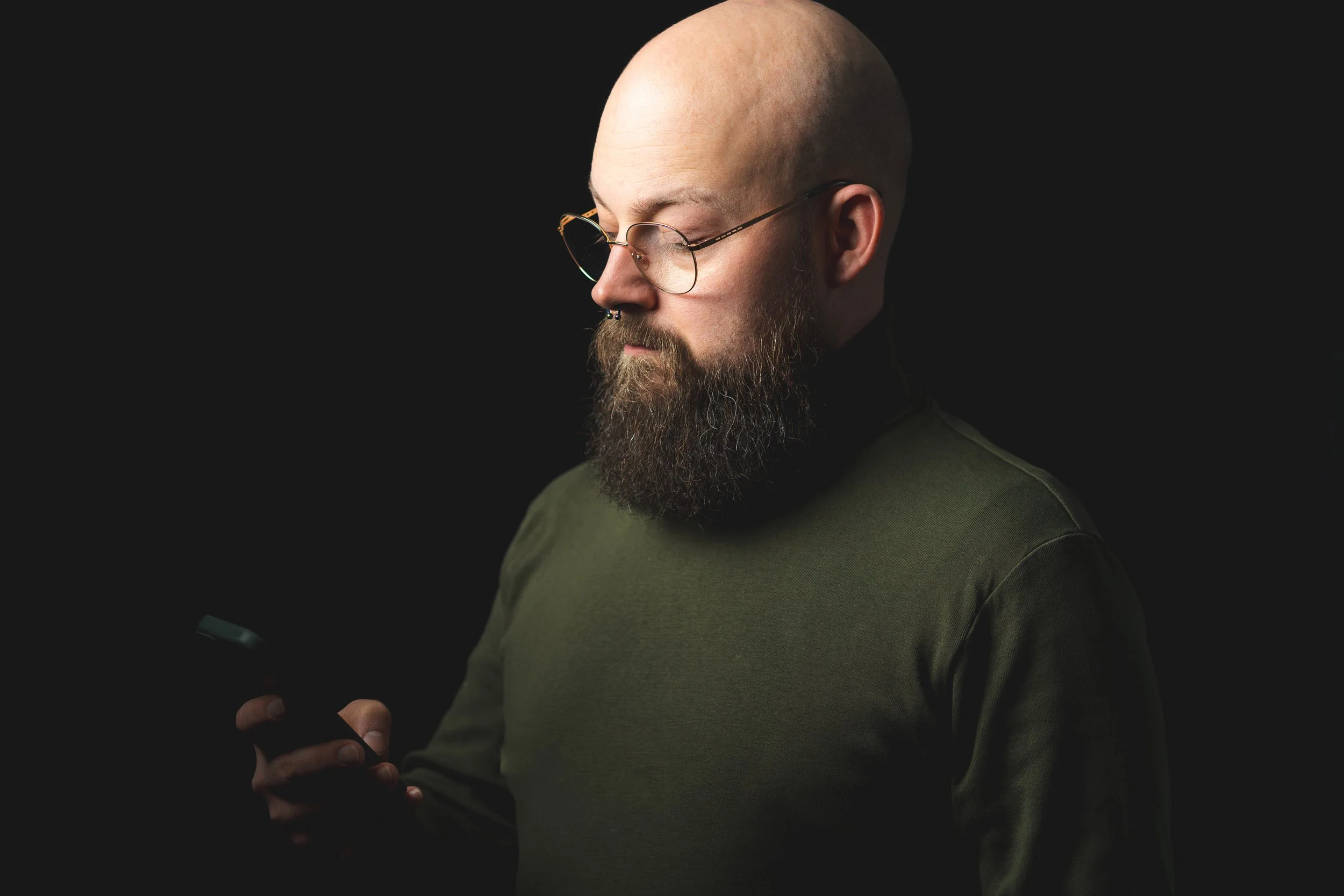 Mann mit Brille, Vollbart und Glatze, trägt einen dunkelgrünen Pullover, schaut auf ein Smartphone in der Hand, dunkler Hintergrund.