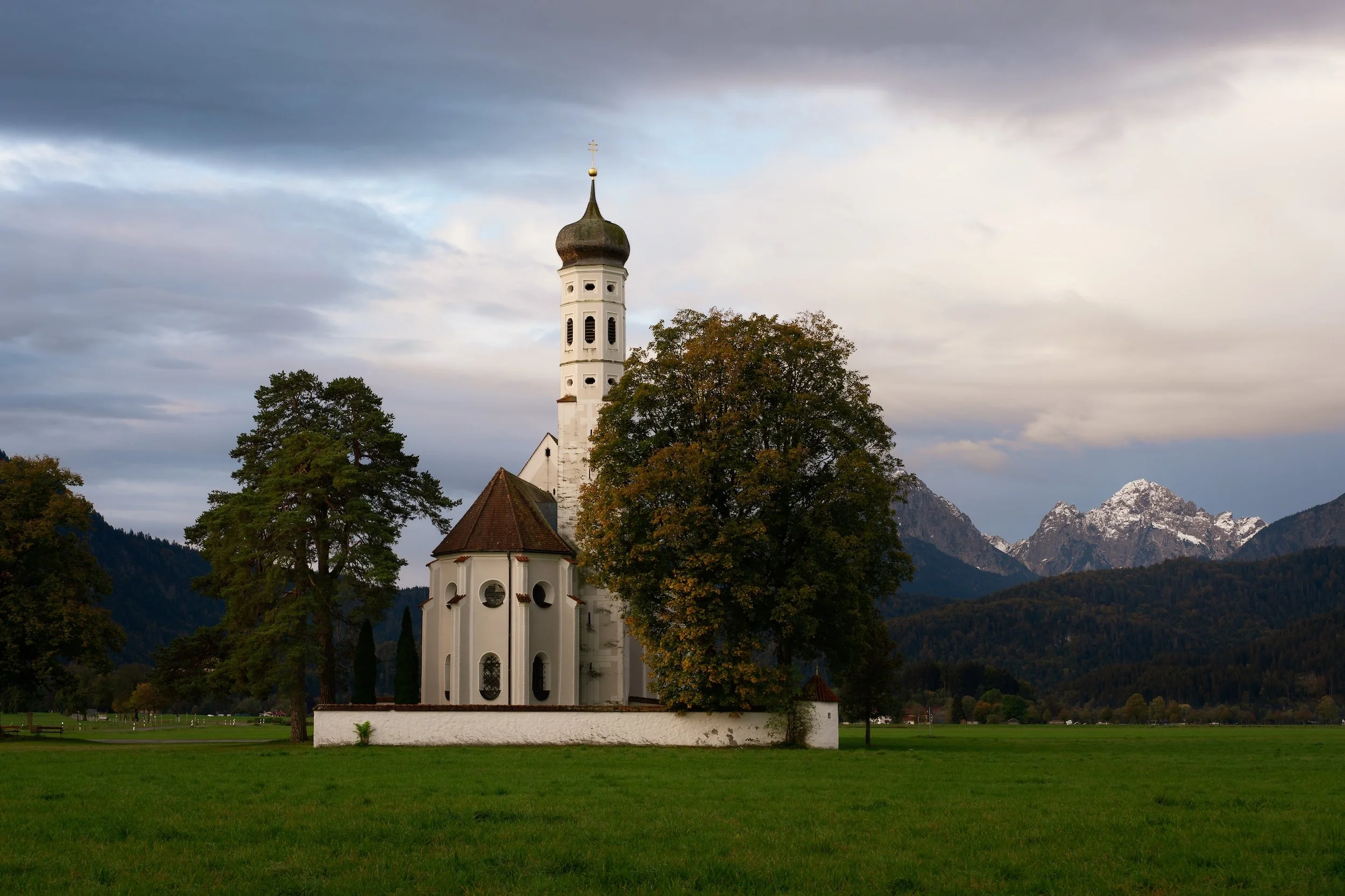 Eine weiße Kirche mit dunklem Turm in einer grünen Wiese, umgeben von Bäumen, mit Bergen im Hintergrund unter einem bewölkten Himmel.