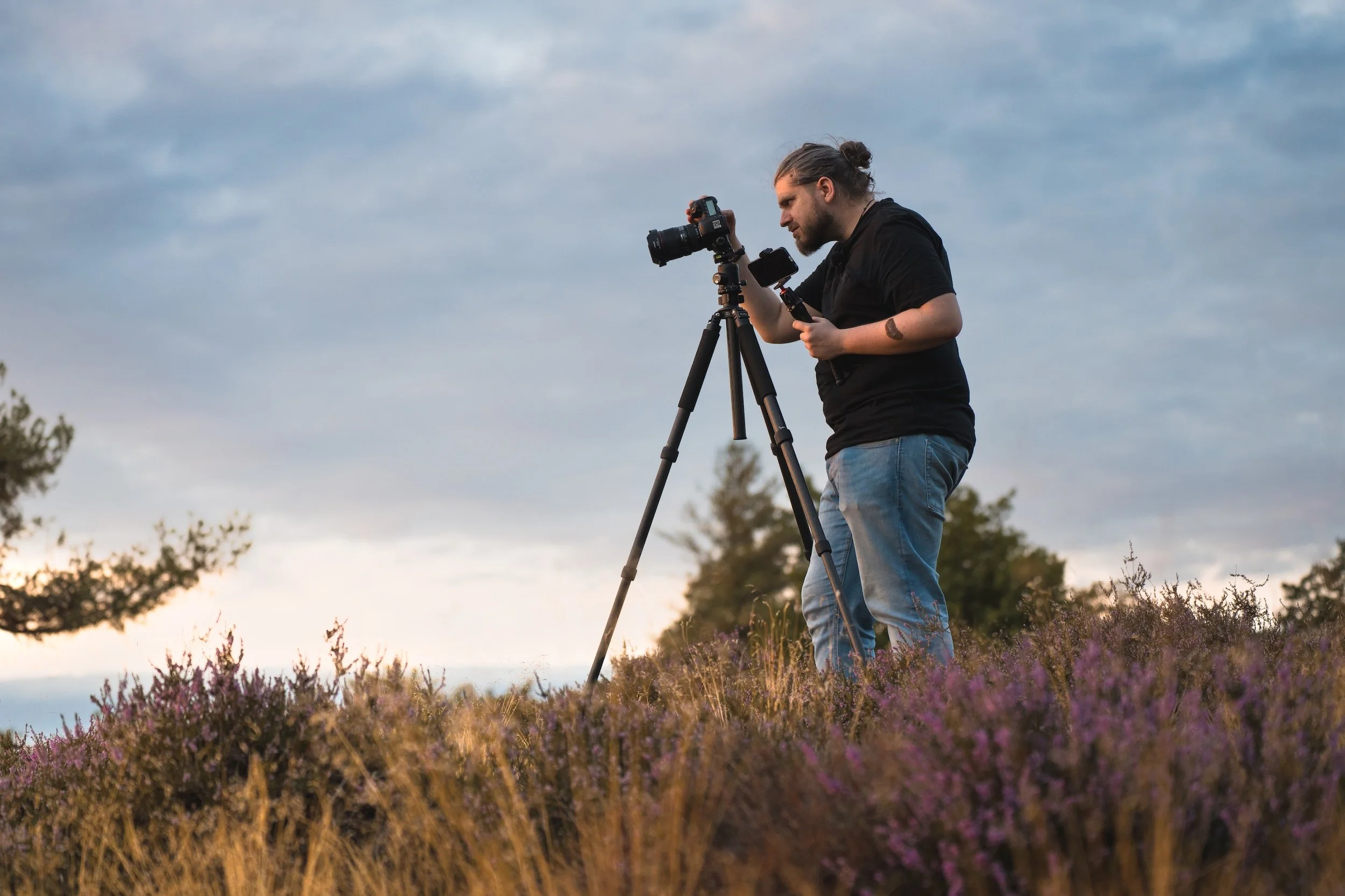 Ein junger Mann fotografiert mit Kamera auf Stativ in einer ländlichen Blumenwiese bei Sonnenuntergang.