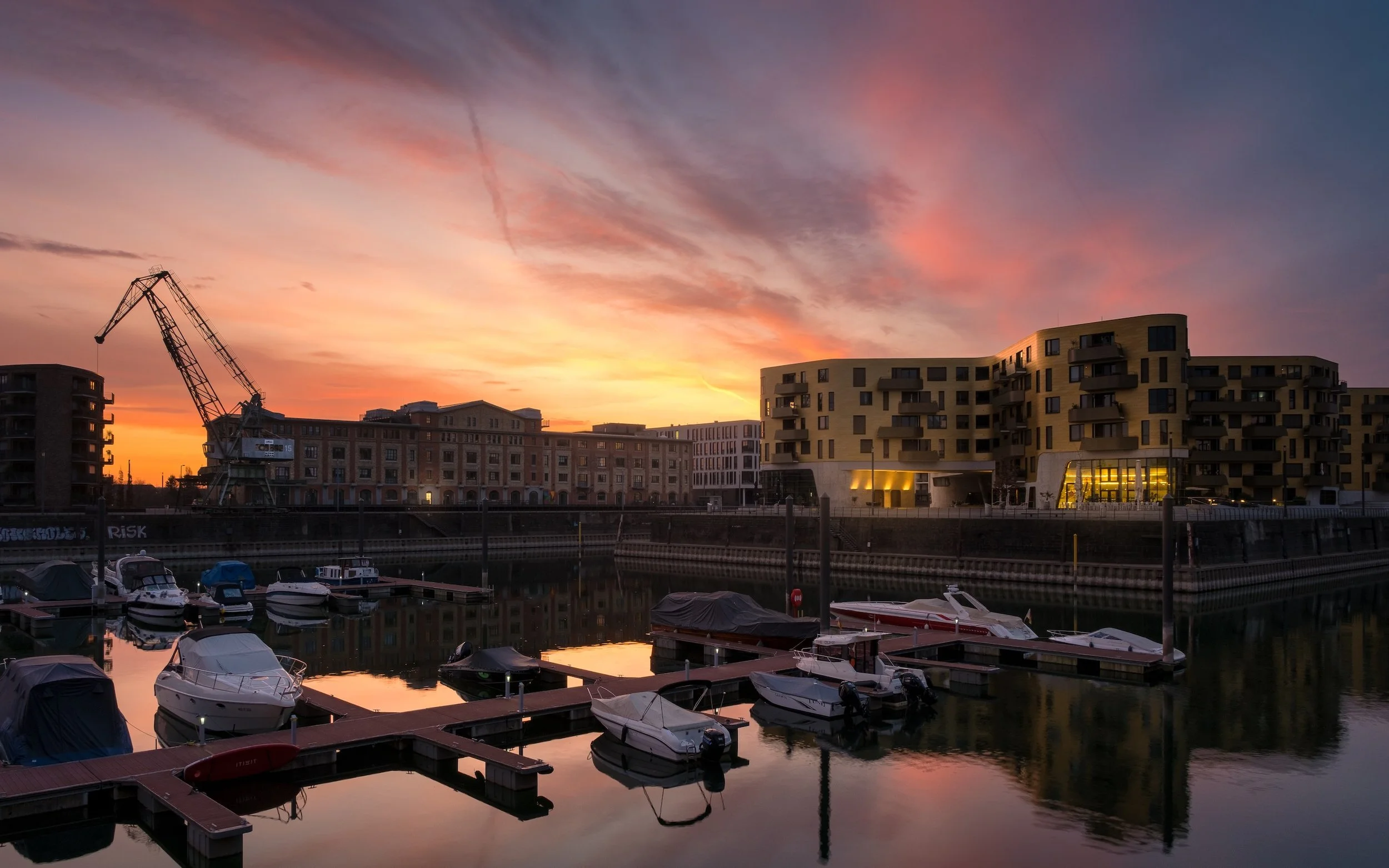Ein Hafen mit Booten am Wasser bei Sonnenuntergang, moderne Gebäude im Hintergrund, der Himmel ist in Orangetönen mit Wolken