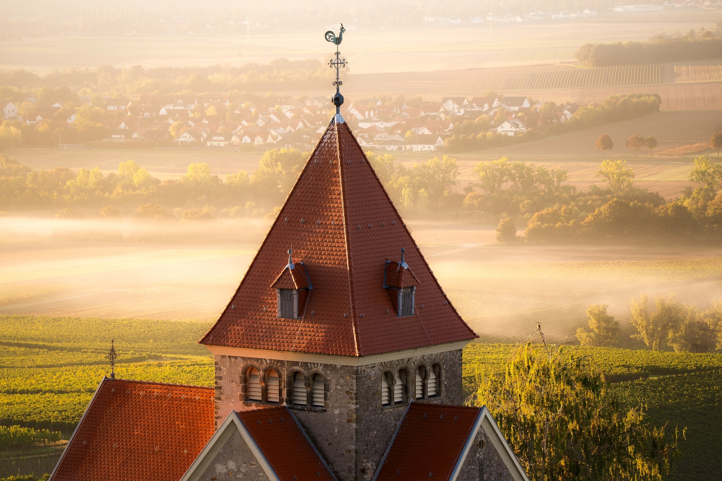 Ein Kirchturm mit roten Ziegeldächern vor einem nebelbedeckten ländlichen Panorama mit Feldern, Bäumen und einem Wäldchen bei Sonnenaufgang.