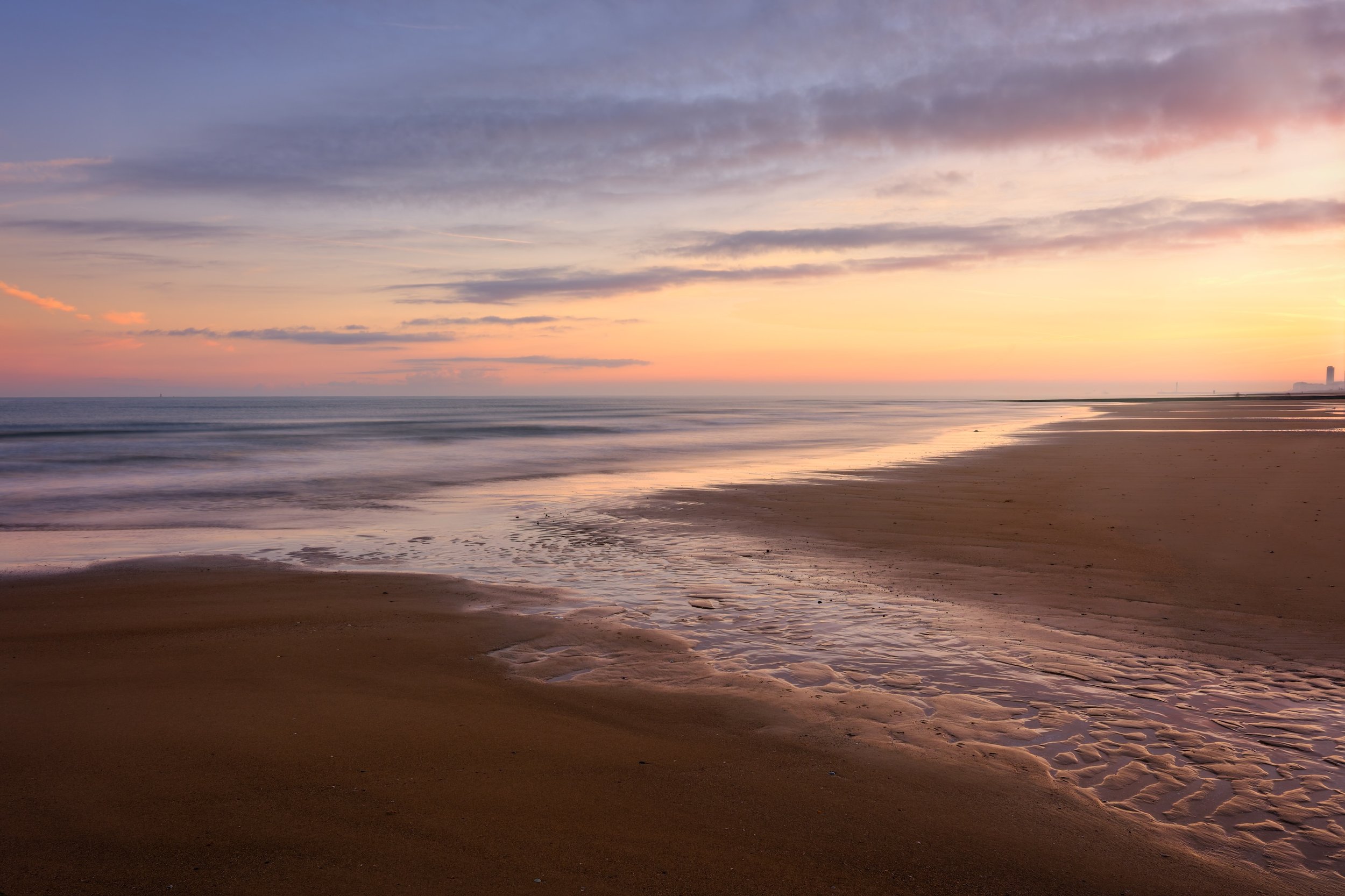 Ein ruhiger Strand bei Sonnenuntergang mit weichem Himmel in Pastellfarben und Wellen, die sanft an das Ufer schlagen.