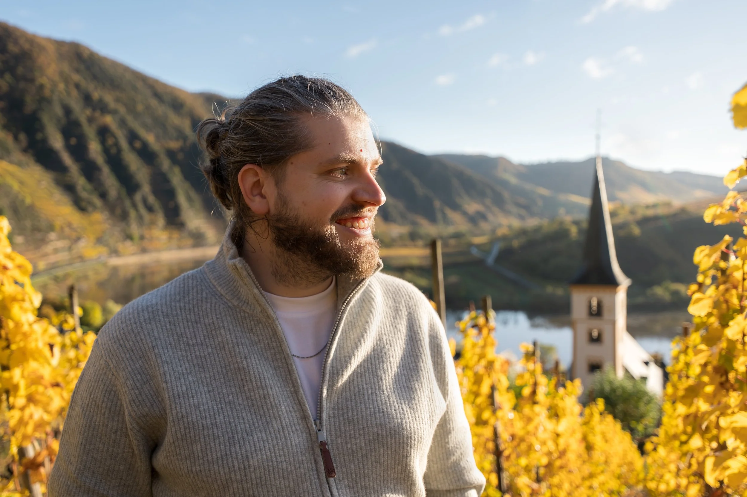 Junge Mann mit Bart und langen Haaren in einem hellgrauen Pullover, lachend im Weingarten bei Sonnenuntergang, mit Kirche und Bergen im Hintergrund.