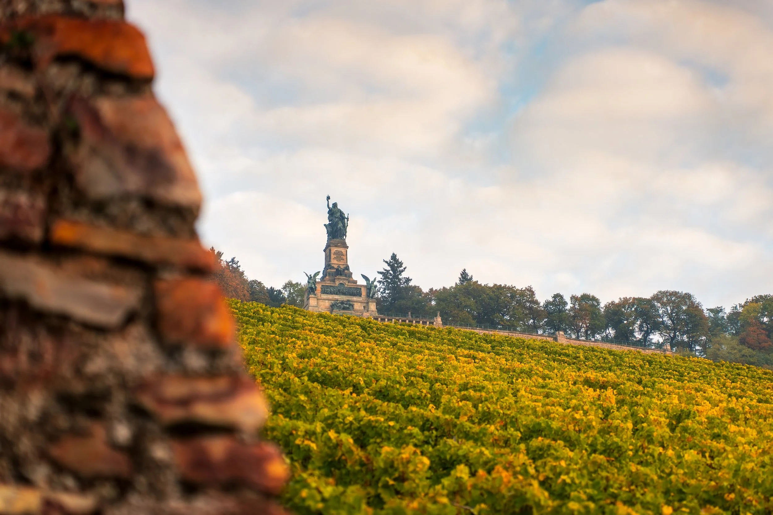 Fokus auf eine antike Ziegelmauer im Vordergrund, während im Hintergrund eine Statue auf einem Hügel mit Weinreben und Bäumen zu sehen ist.
