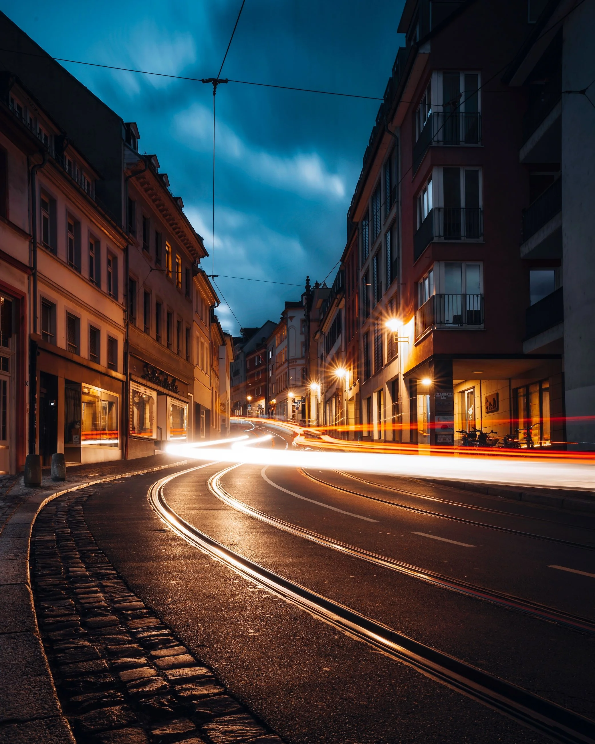 Blick auf eine kurvige Straße in einer Stadt bei Nacht, mit Lichtspuren von vorbeifahrenden Fahrzeugen und beleuchteten Gebäuden an der Seite.