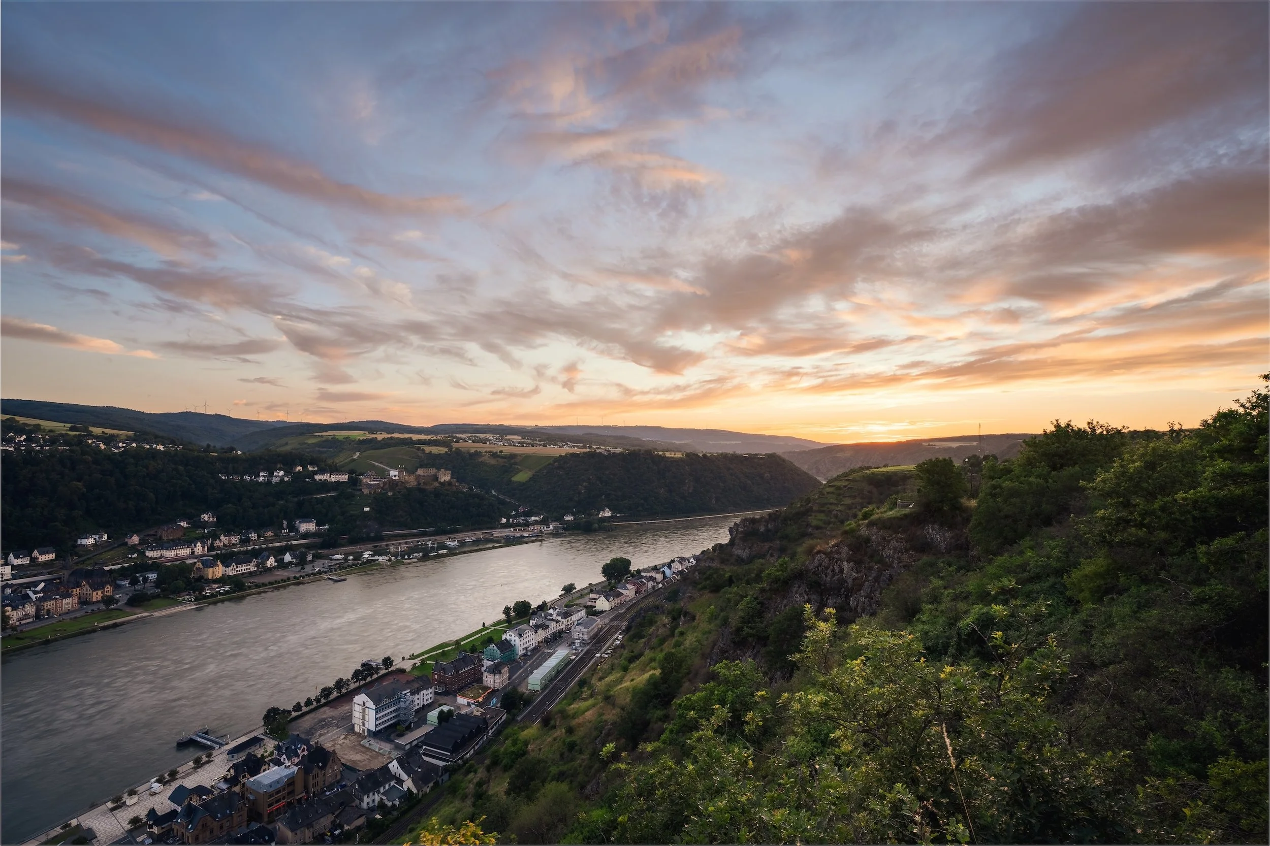 Blick auf den Fluss bei Sonnenuntergang mit Bergen und einer Stadt am Ufer, umgeben von grünen Hügeln und bewölktem Himmel.