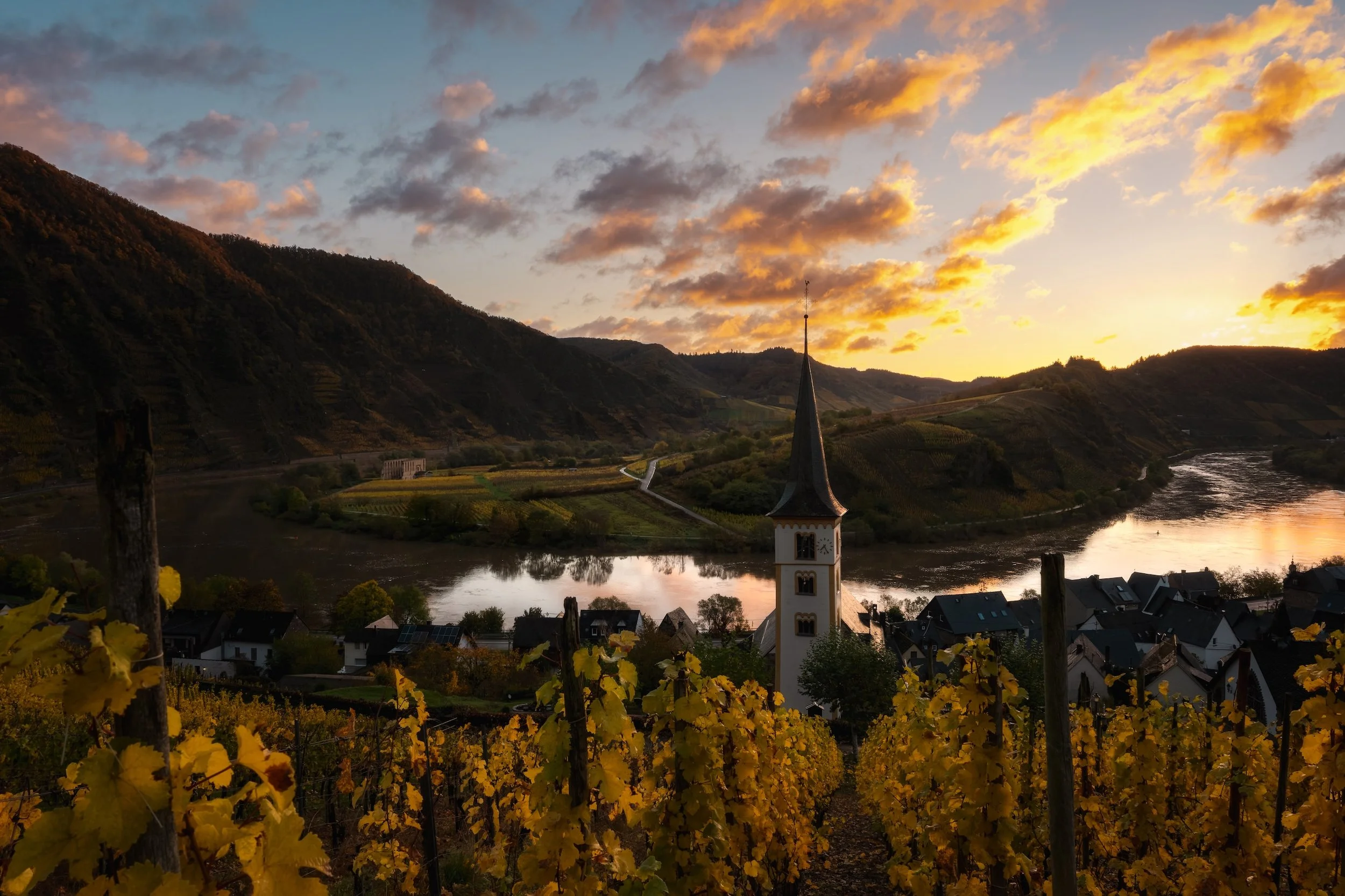 Ein Sonnenuntergang über einem Fluss, umgeben von Hügeln und Weinbergen, mit einer katholischen Kirche im Vordergrund.