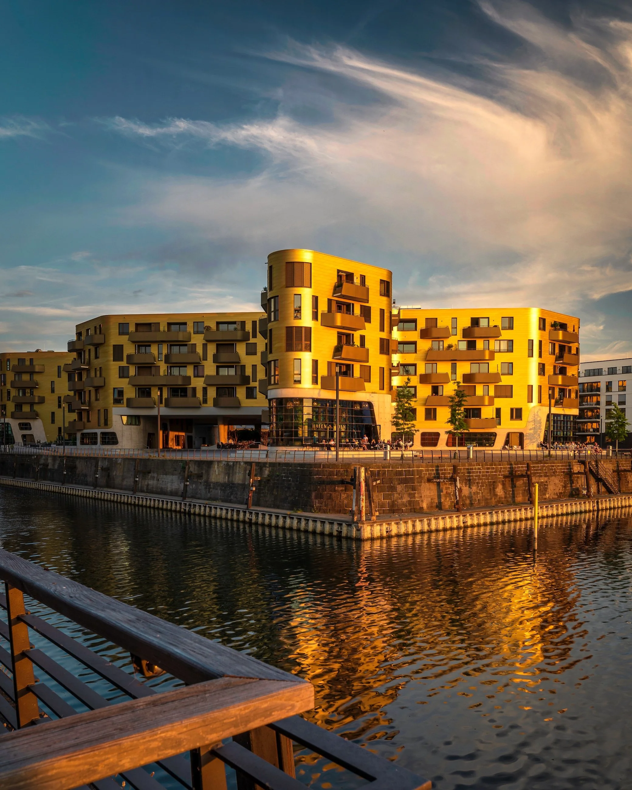 Ein modernes gelbes Gebäude an einem Wasserübergang bei Sonnenuntergang, mit reflektierendem Wasser im Vordergrund und einem Himmel mit leichten Wolken.