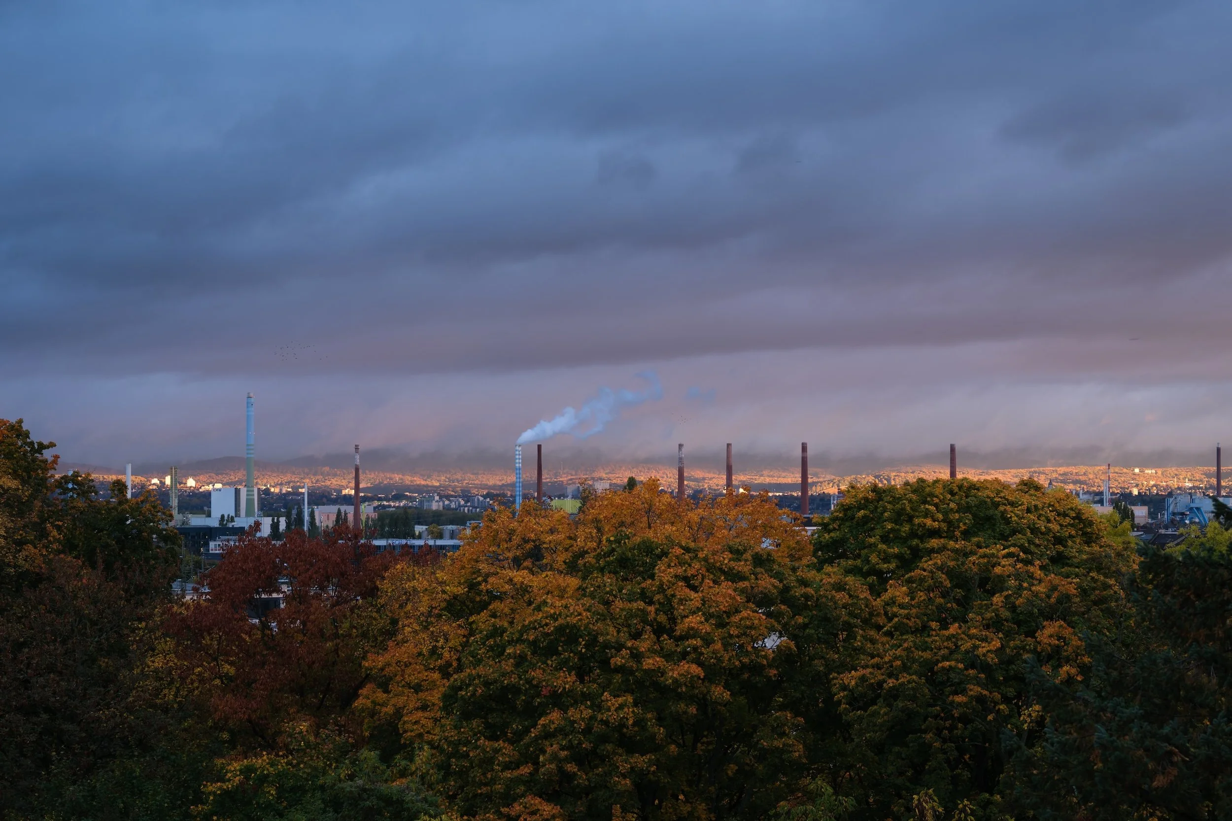 Stadtansicht mit Fabrikschloten, Rauch und herbstlichen Bäumen im Vordergrund, dunkle Wolken am Himmel.