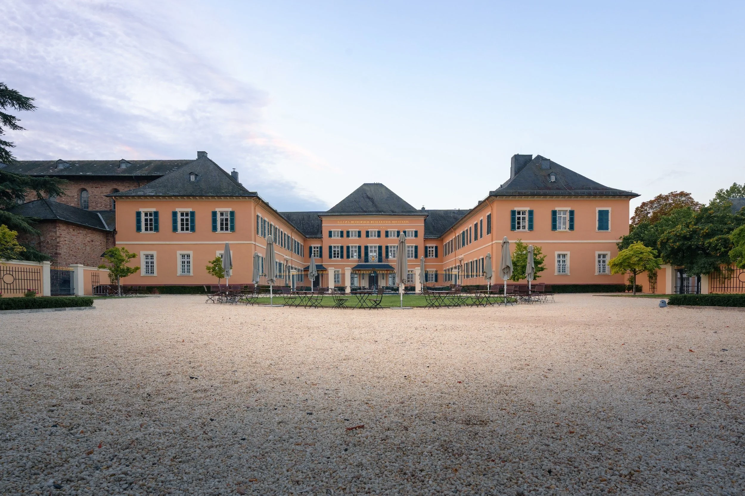 Ein großes, gelb-orangefarbenes Gebäude mit blauen Fensterläden auf einem gepflegten Gelände mit Tischen und Schirmen im Vordergrund.
