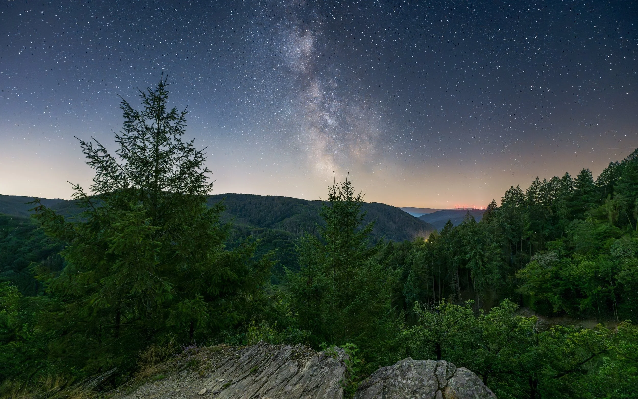 Nachtaufnahme eines Sternenhimmels mit sichtbarer Milchstraße über einem bewaldeten Hügel mit Felsen im Vordergrund.
