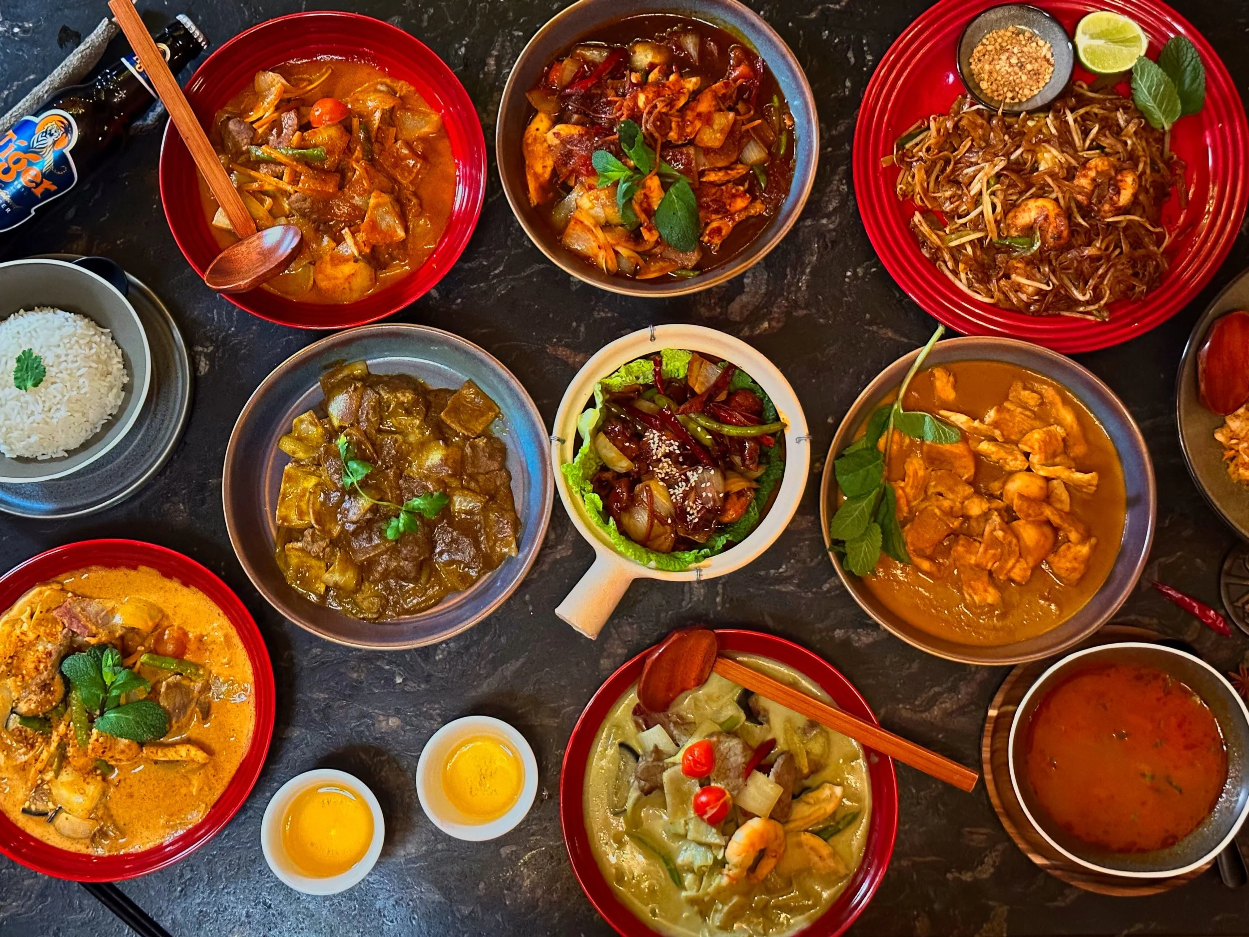 Assorted Thai dishes on a dark table, including various curries, stir-fries, rice, noodle dishes, and condiments with lime and herbs.