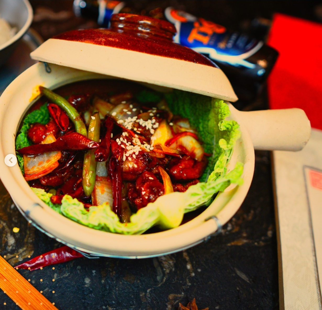 A ceramic mug with a lid, filled with spicy Asian-style stir-fry vegetables and meat, garnished with sesame seeds, on a dark surface.