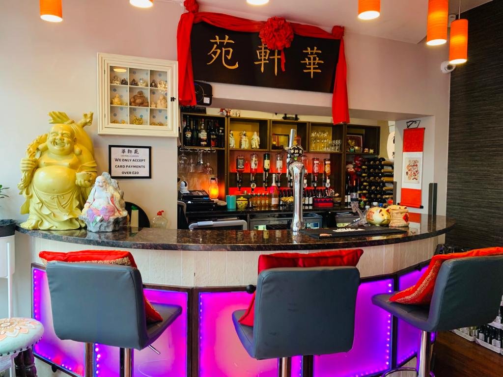 A bar with a curved counter illuminated with purple lights, three gray bar stools with red cushions, and Asian decorations including a large yellow laughing Buddha statue and a smaller figurine, a black and gold Chinese character sign with red fabric, and a back wall with bottles of alcohol and glasses.