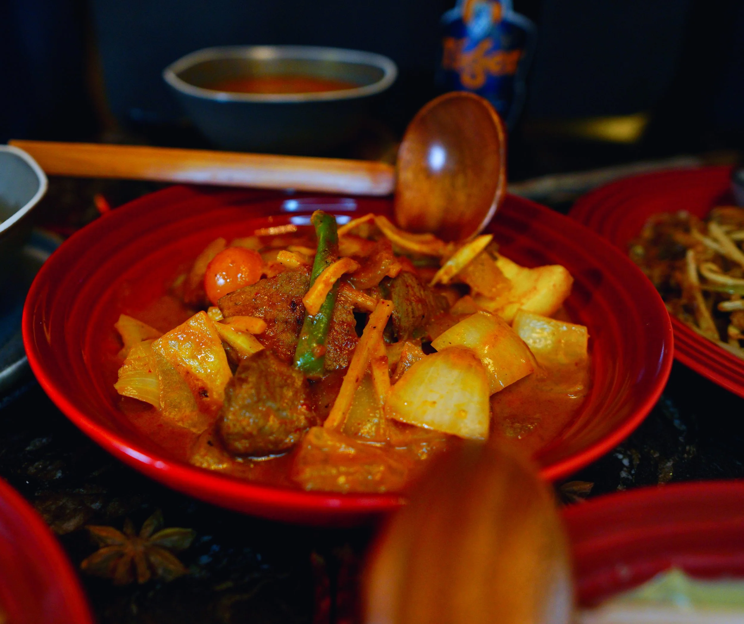 A red bowl filled with Asian-style curry containing beef, vegetables, and potatoes, with a wooden spoon resting inside.