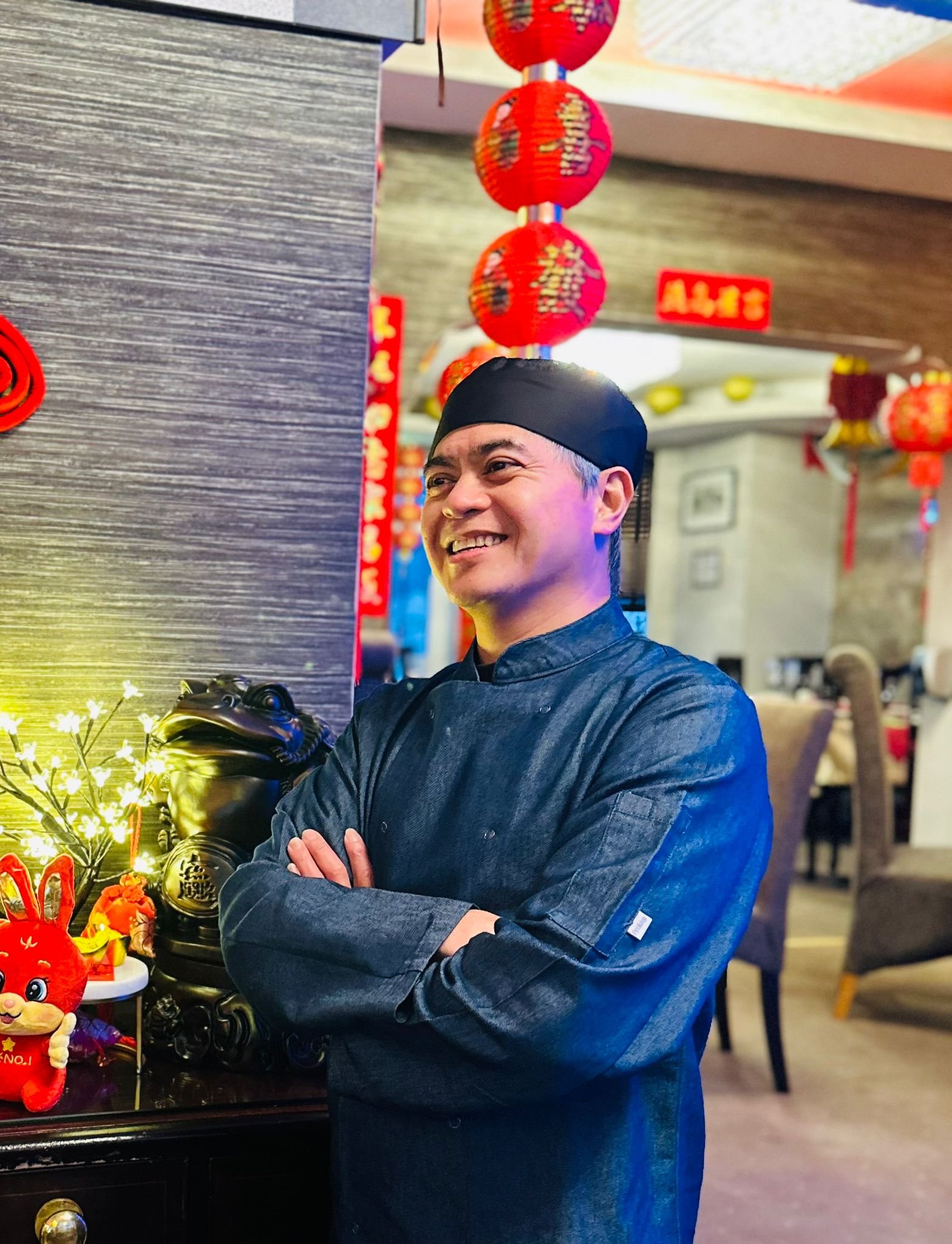 A smiling man in a black chef's coat and black cap standing inside a restaurant decorated with red lanterns and festive decorations.