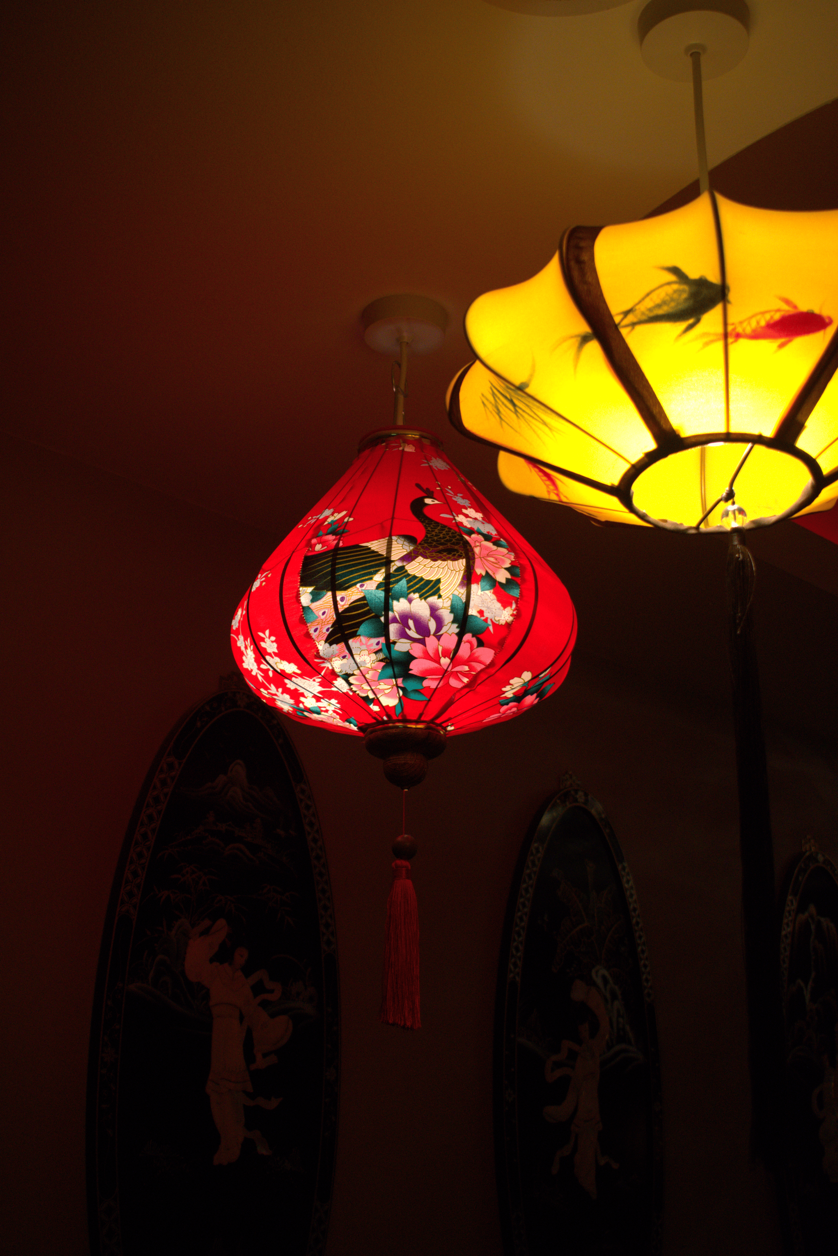Two hanging Asian-style paper lanterns, one red with a peacock and floral design, and one yellow with a floral design, illuminated in a dark room.