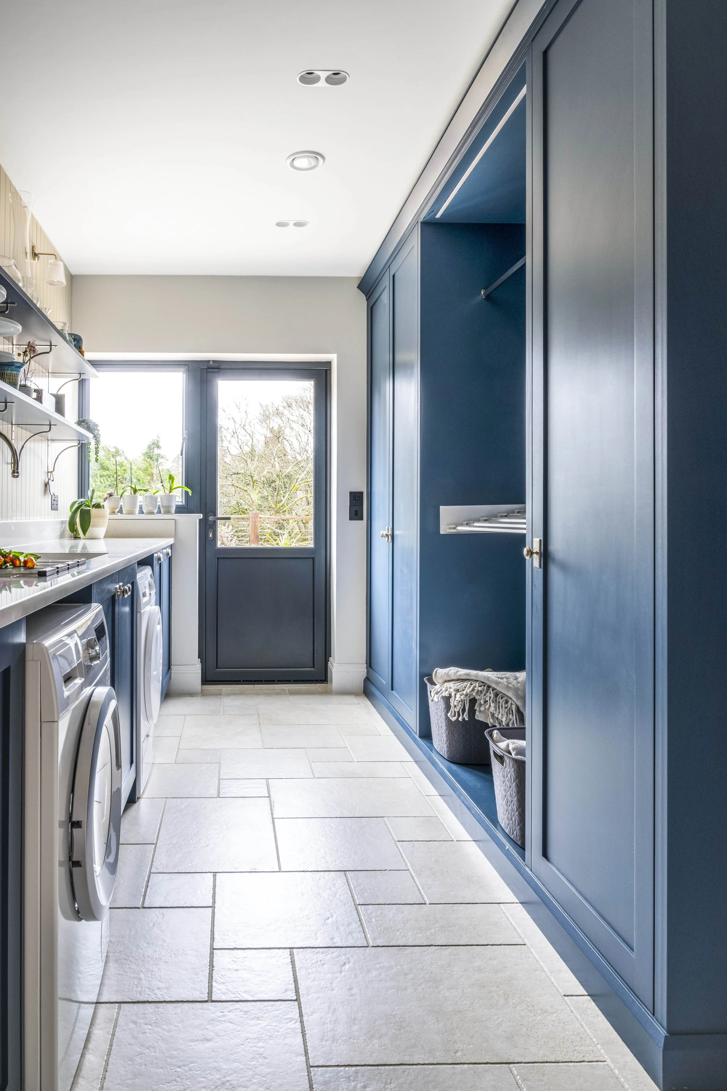 Bright laundry room with white tile floor, blue cabinets, a door leading outside, and laundry appliances along the left wall.