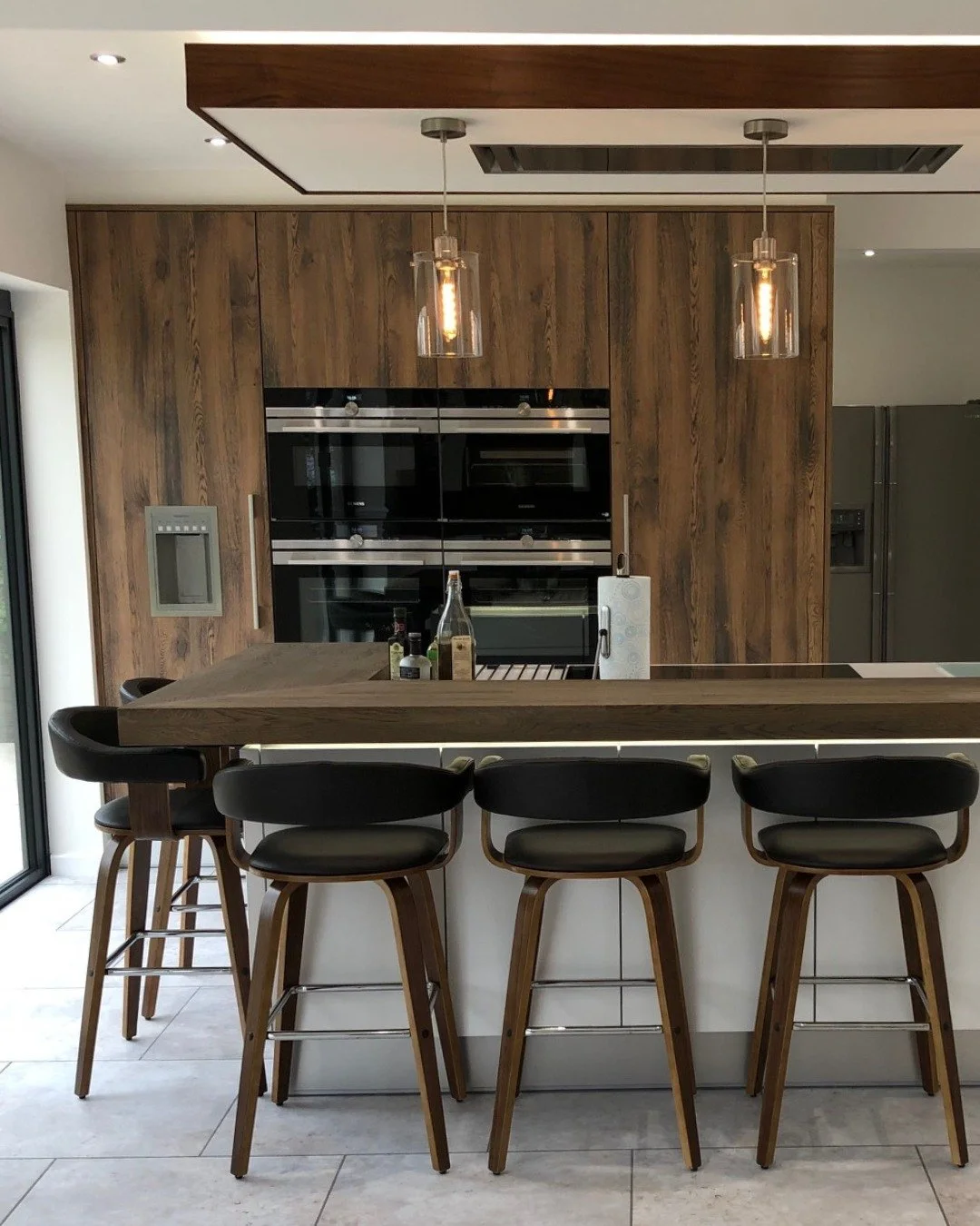 Dark wood grain cabinetry, leather-effect bar stools and a Siemens tower that disappears into the run.

This kitchen was designed around a client who entertains seriously - a layout that separates the cooking from the socialising, with a bar counter 