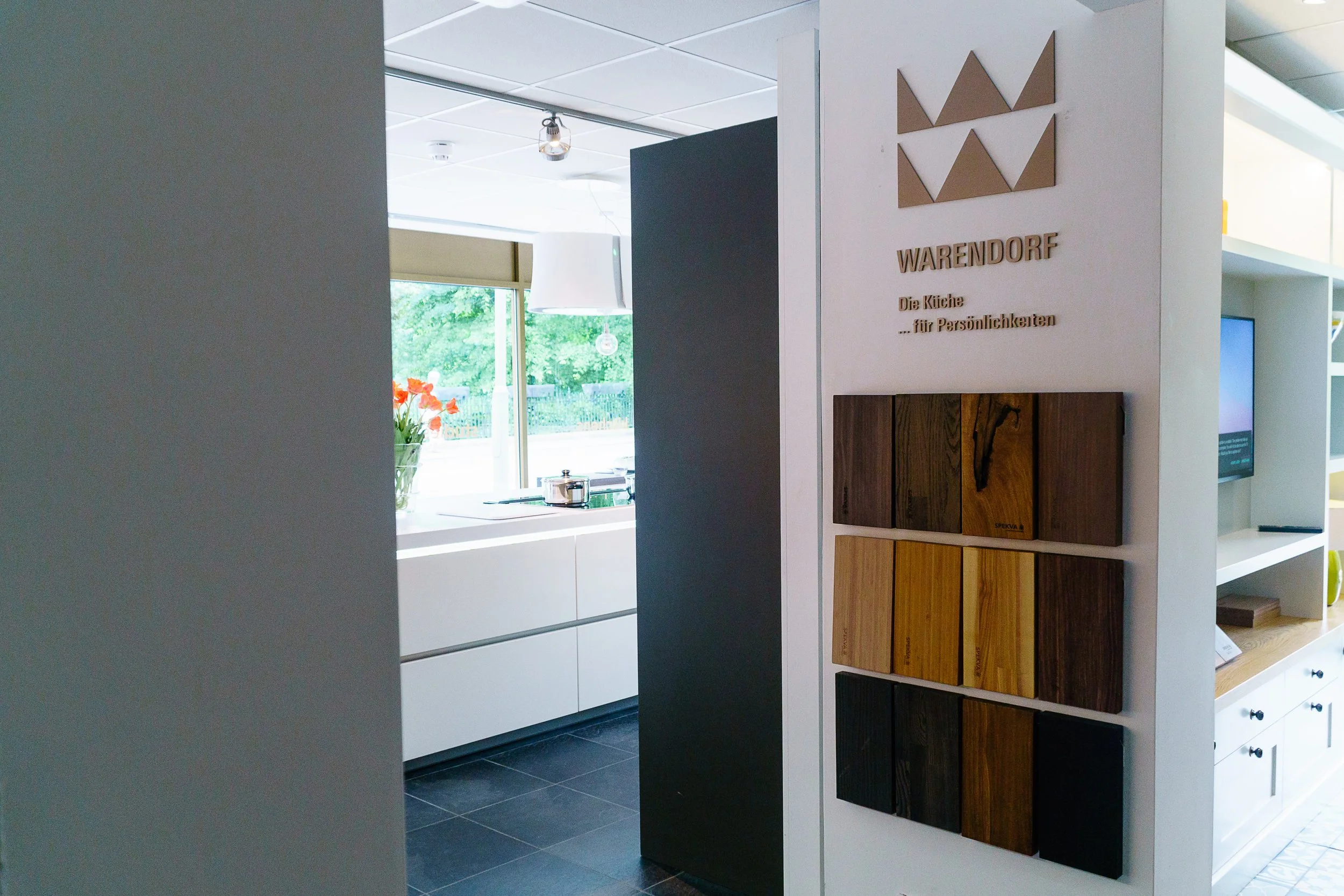 Interior view of a modern kitchen showroom with a display of wooden cabinet samples on a white wall, a background of a kitchen with white cabinets, a window revealing greenery outside, and a vase with orange flowers on the counter.