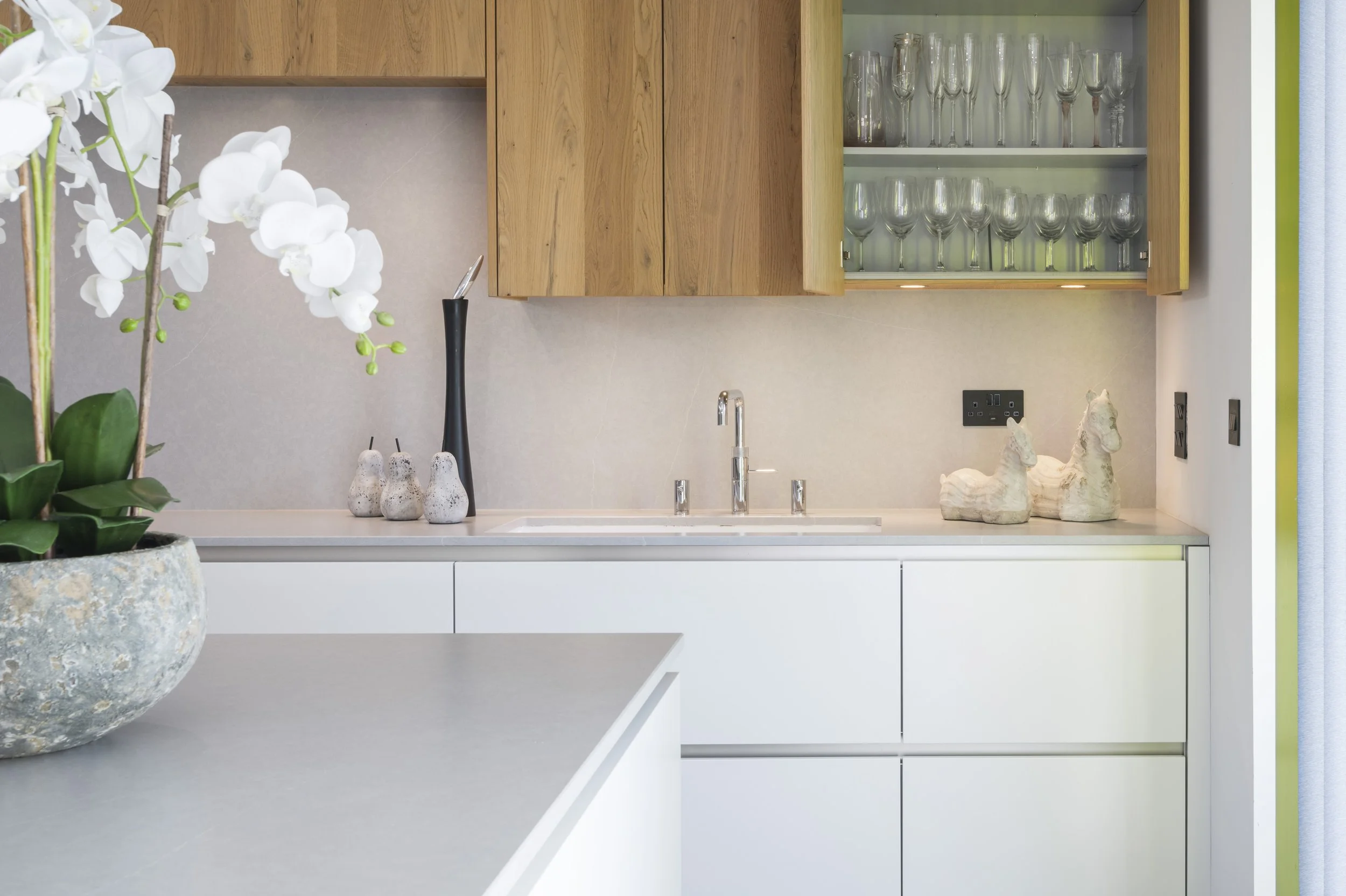 Modern kitchen with white cabinets, a grey countertop, wooden upper cabinets, and a glass-front cabinet with glassware. Decor includes white orchid flowers, small decorative objects, and horse figurines.