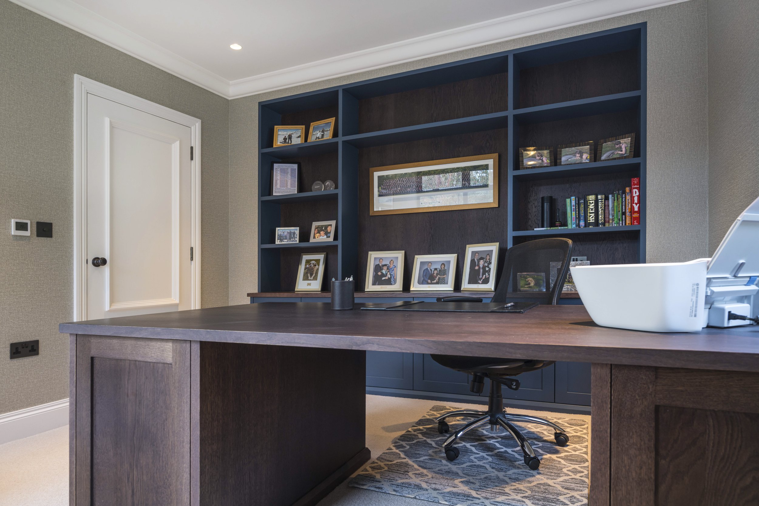 Home office with a large wooden desk, black office chair, blue bookshelf filled with framed photos, books, and decorative items, white door, and beige walls.