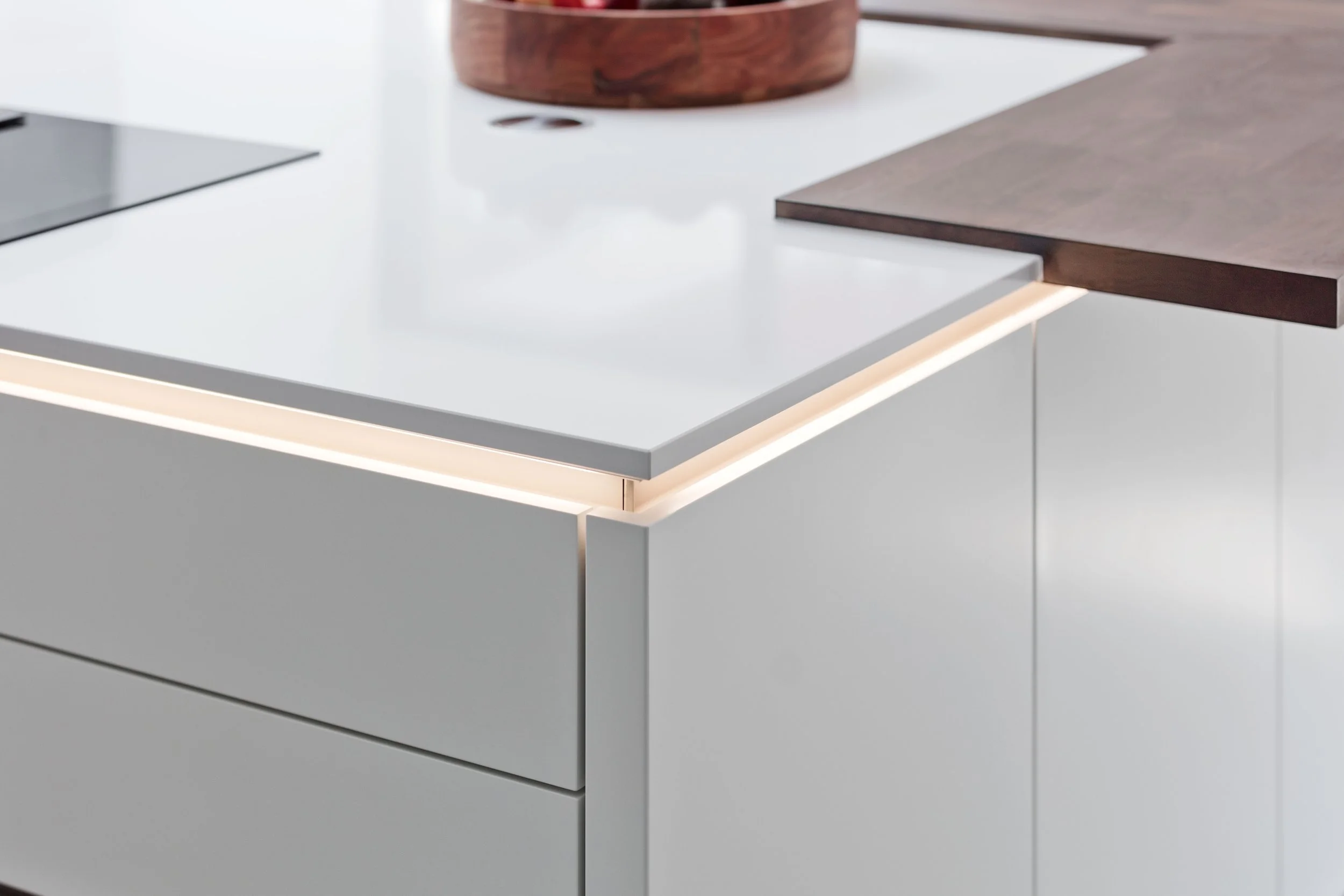 Close-up of a modern kitchen with white cabinets, a black cooktop, and a dark wooden countertop with a rounded wooden bowl on it.