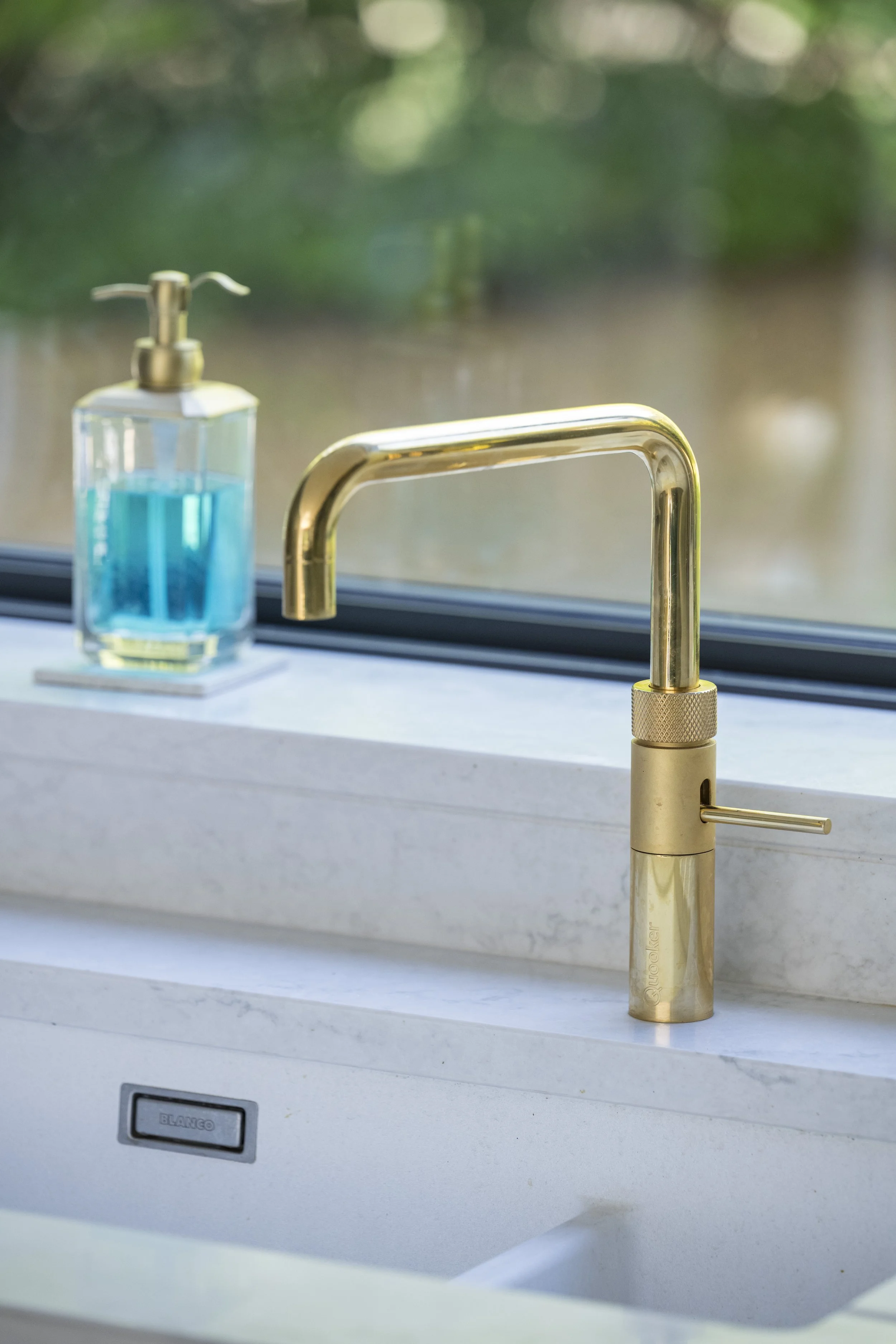 A modern gold kitchen faucet with a bottle of blue liquid soap in a clear container with a gold pump, set on a white countertop by a window with a blurred outdoor view.