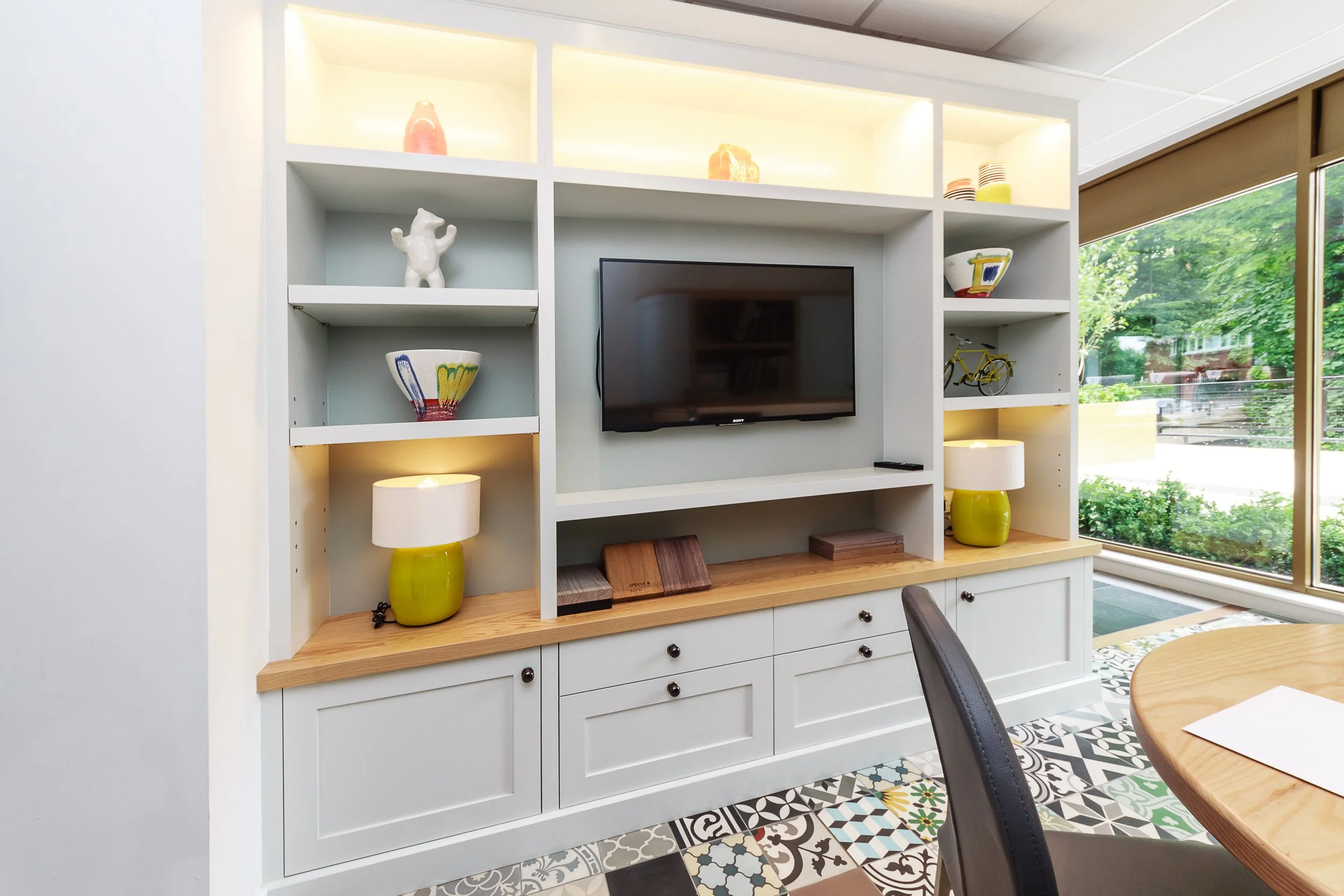 A white entertainment center with open shelves and drawers, yellow lamps, colorful bowls, decorative items including a bicycle figurine, and a flat-screen TV in a room with large windows and patterned tile flooring.
